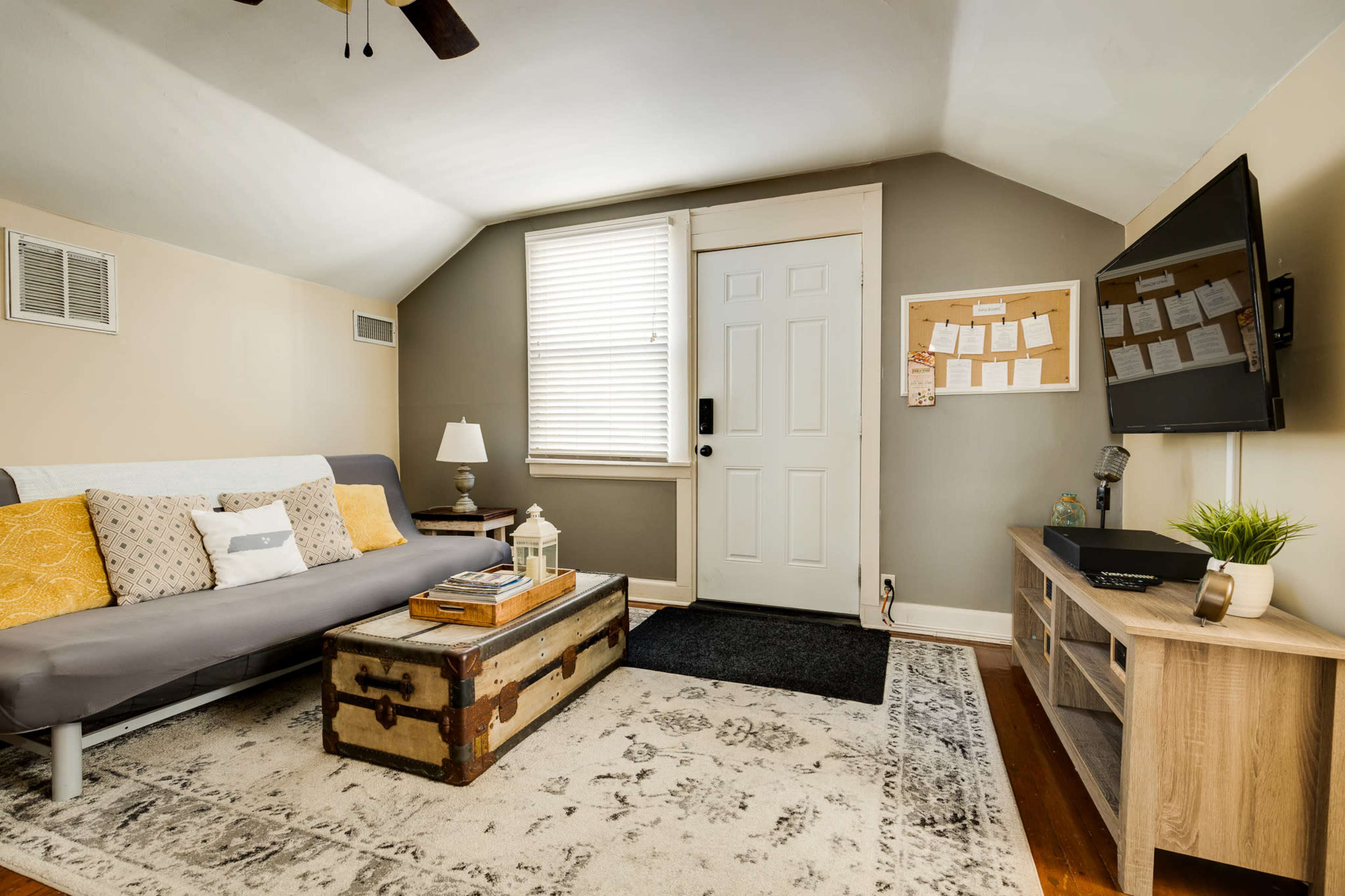 The image shows a cozy living room with a gray couch, a wooden coffee table, a television mounted on the wall, and a door leading outside.
