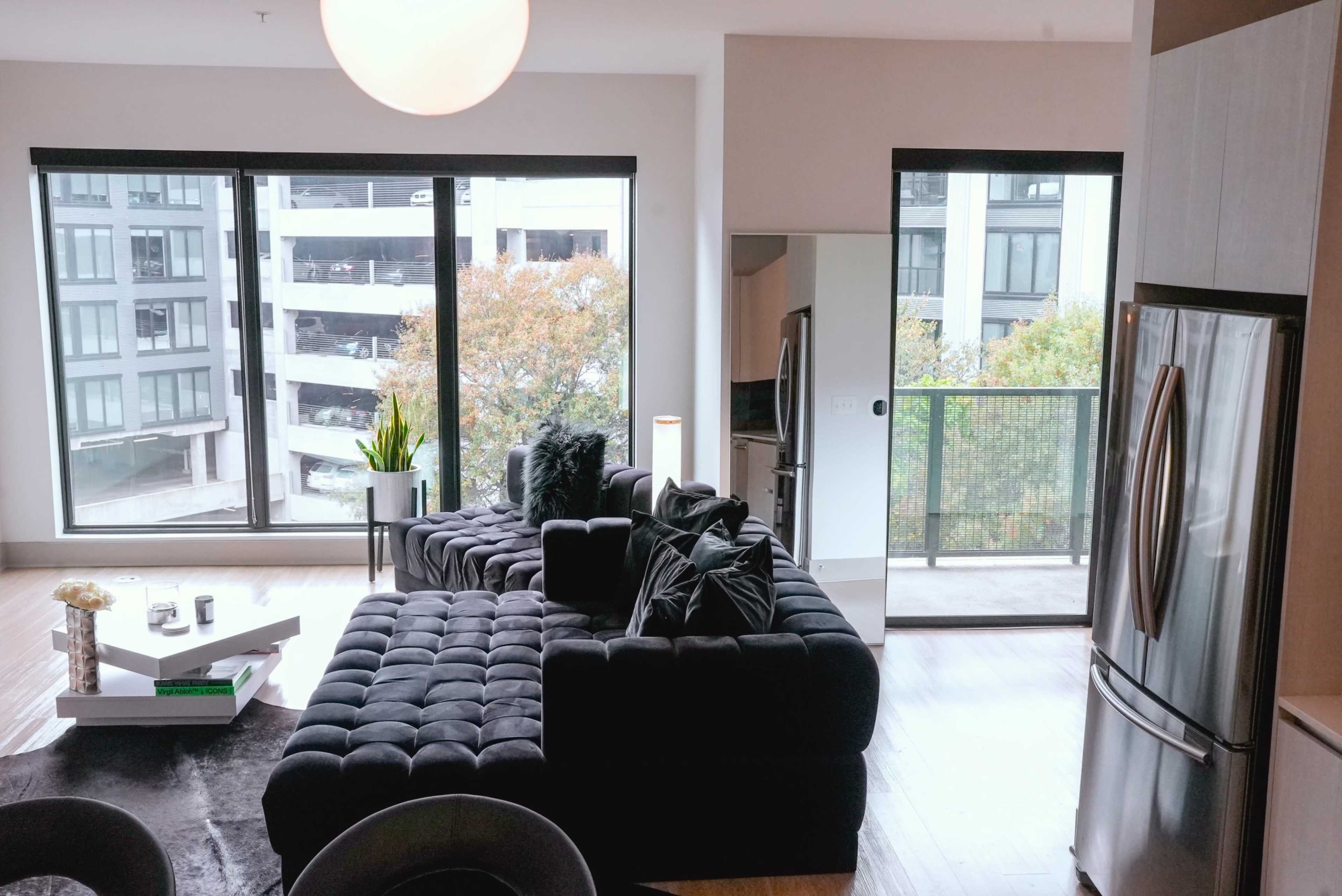A modern living room features a black sectional sofa, large windows overlooking a balcony, and sleek kitchen appliances.