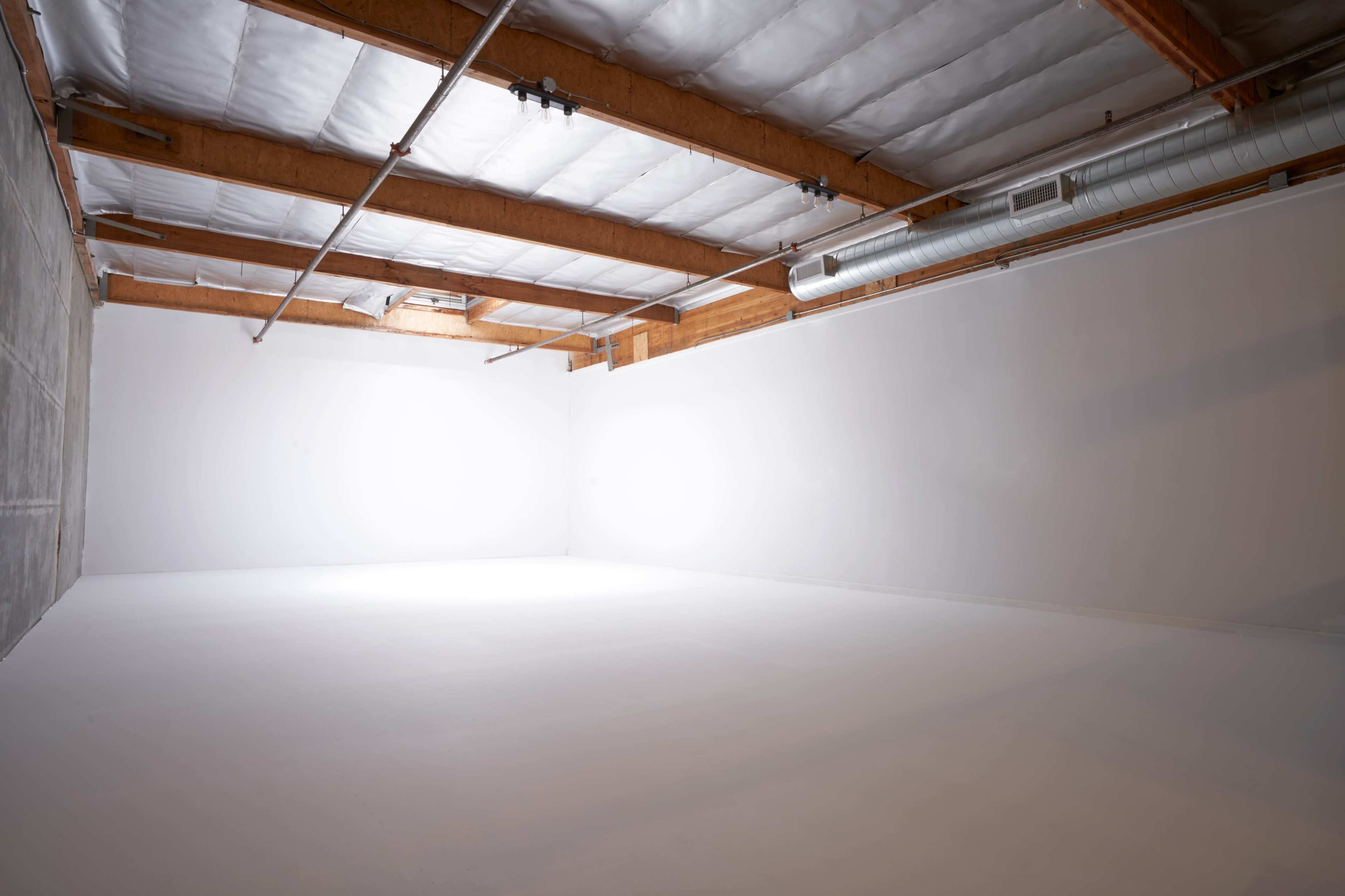 A spacious, empty room with white walls and a wooden beam ceiling, illuminated by overhead lights.