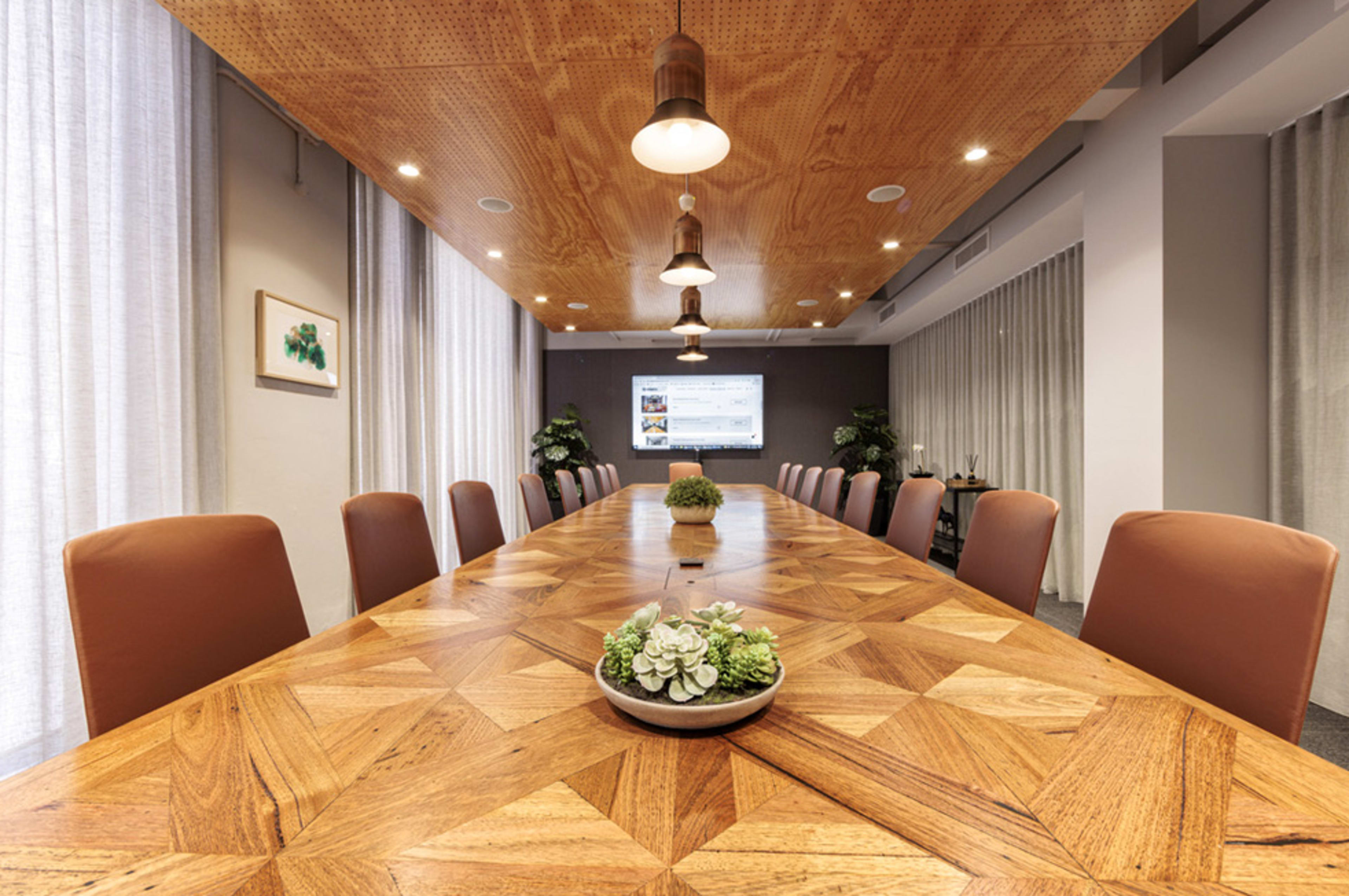 A large conference room features a wooden table with a geometric design, surrounded by brown chairs, and is illuminated by hanging lights with a screen displaying content at one end.