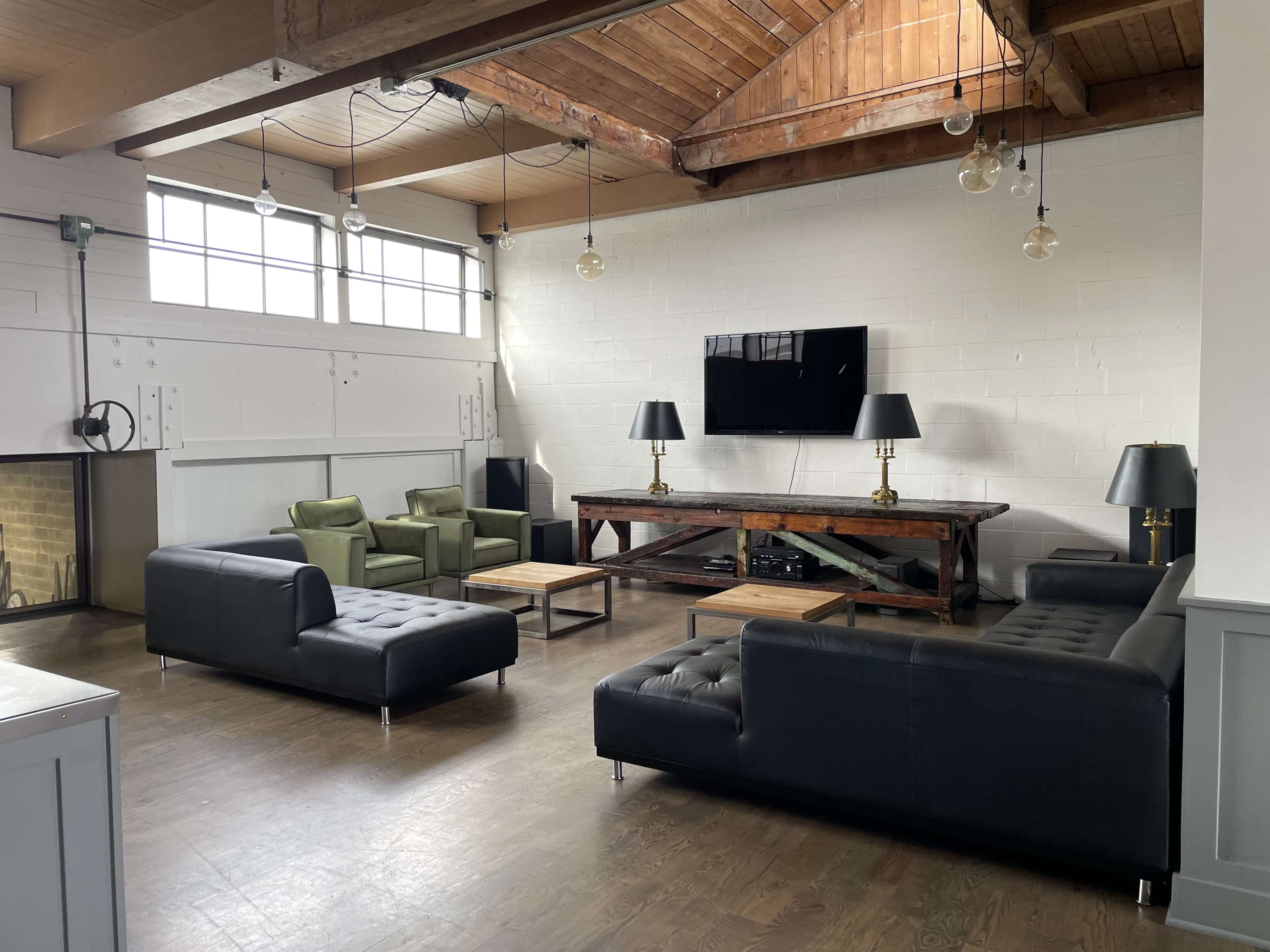 A spacious seating area featuring dark leather sofas, a wooden coffee table, and a wall-mounted television under a high ceiling with visible rafters and hanging light fixtures.