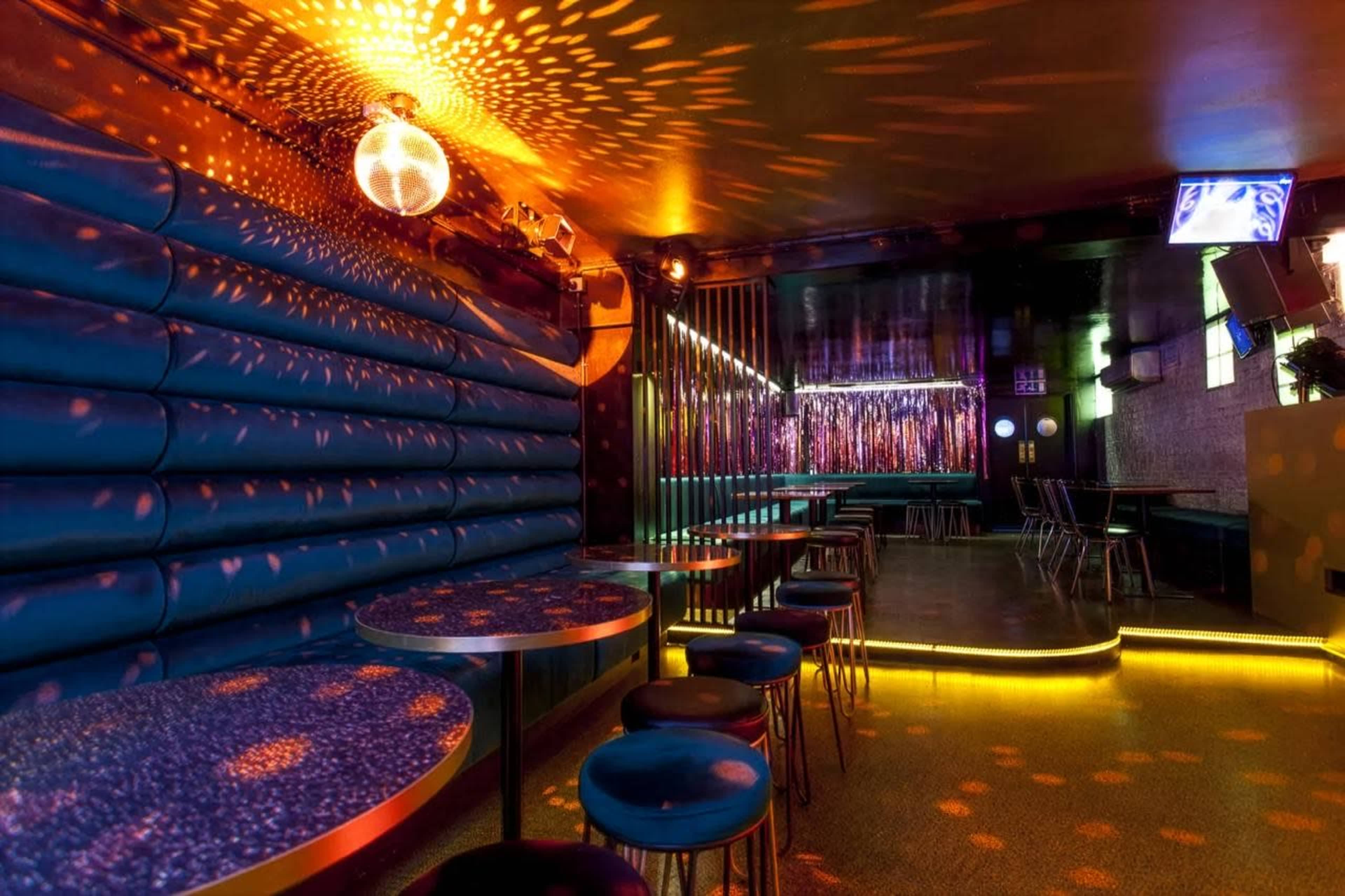 The image shows a dimly lit nightclub interior featuring blue seating, circular tables, and a disco ball reflecting colorful lights across the walls.