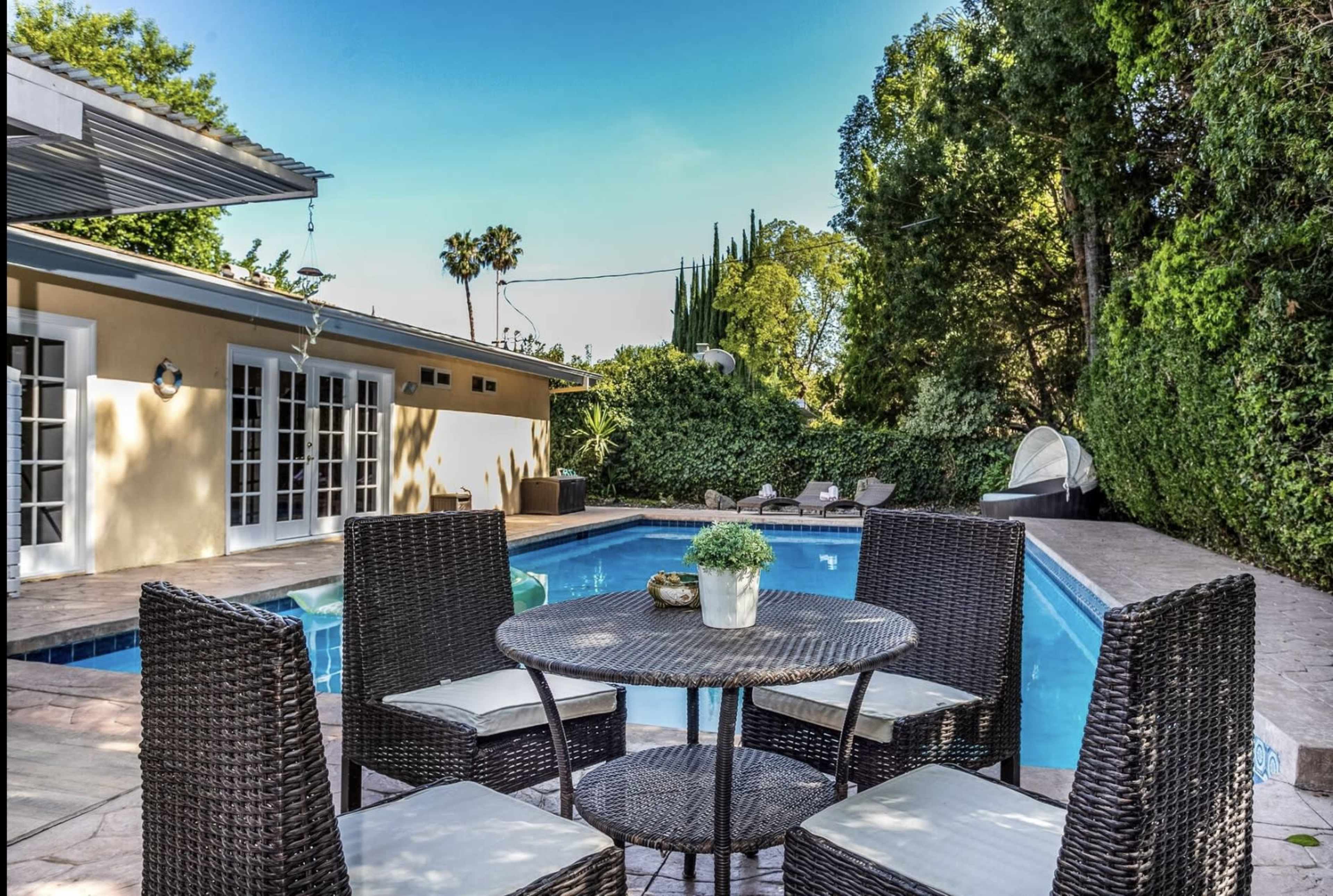 A circular outdoor table with four chairs is set beside a rectangular swimming pool surrounded by greenery and a yellow building in the background.