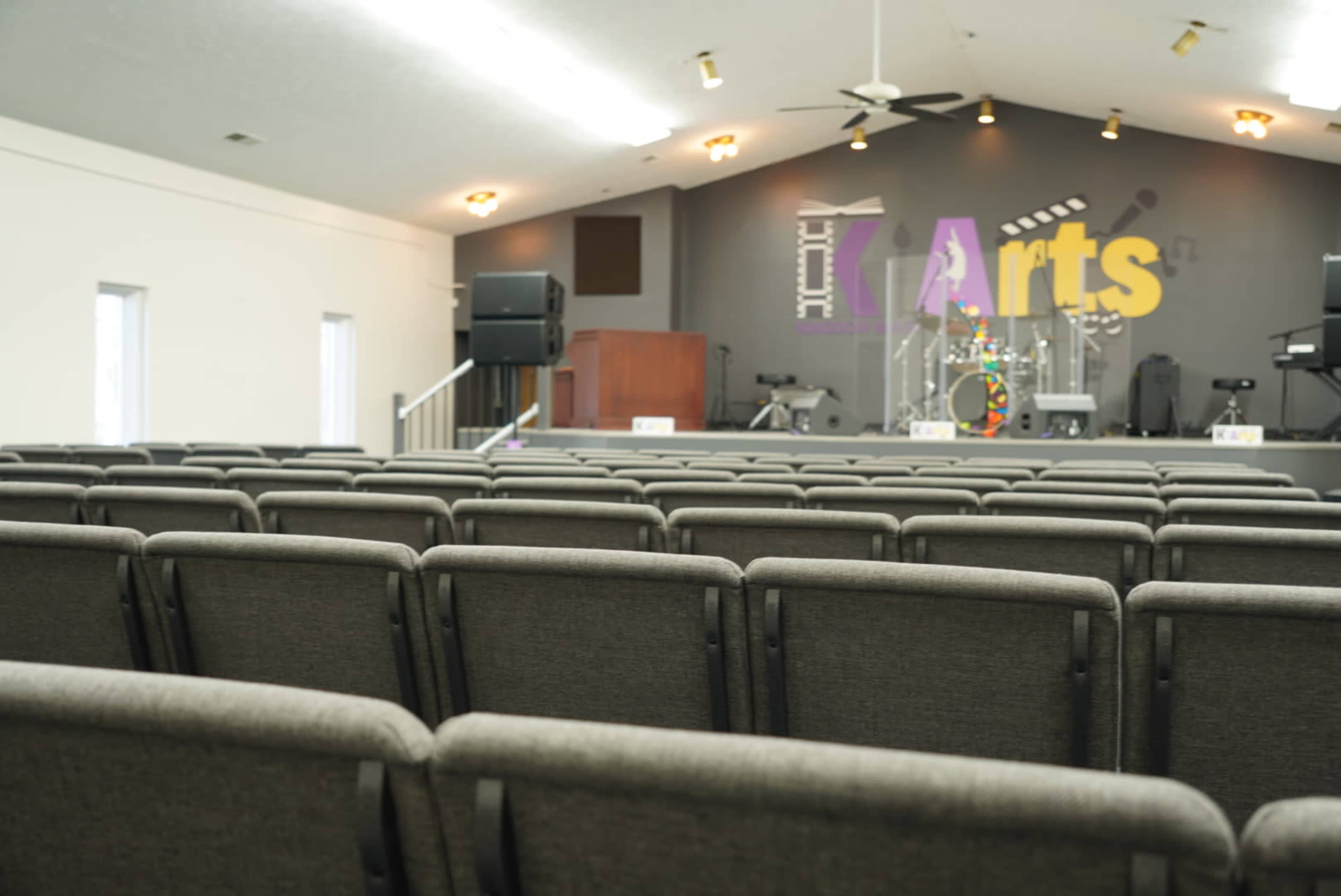The image shows a row of gray chairs arranged in a spacious auditorium with a stage featuring musical instruments and colorful lettering on the wall.