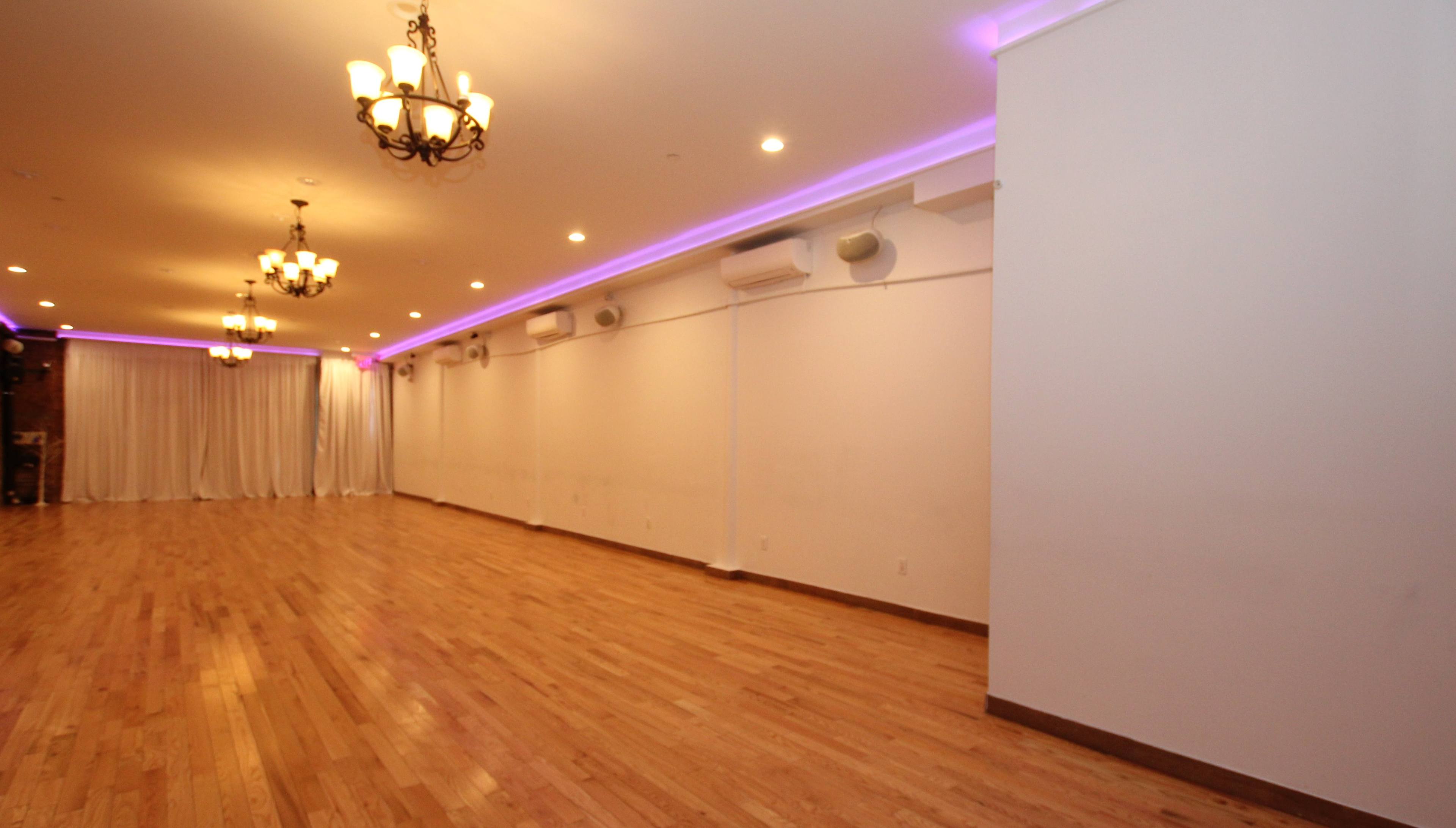 A well-lit event space with wooden flooring, chandeliers hanging from the ceiling, and white drapes along one wall.