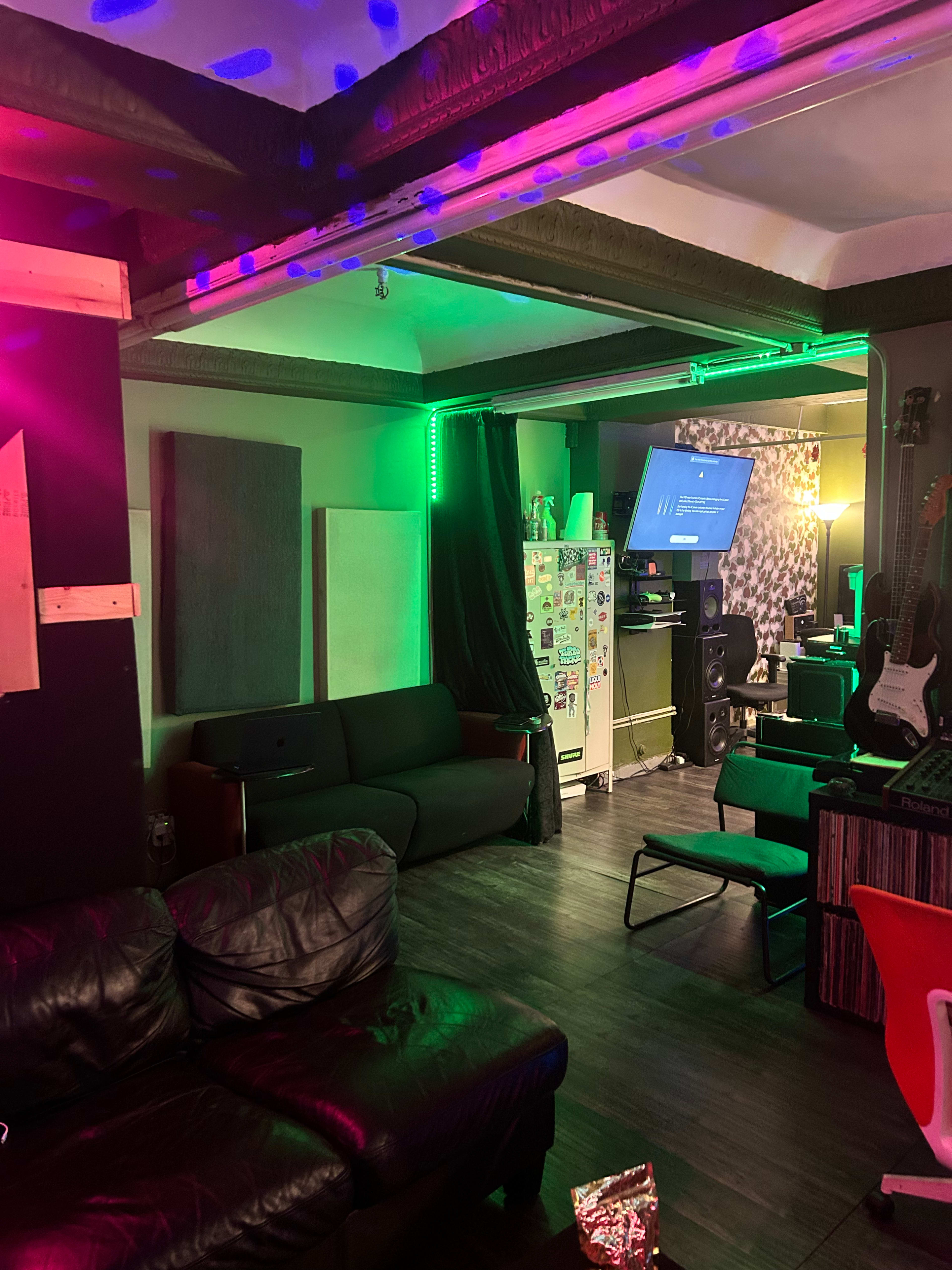 The image shows a dimly lit music studio or lounge area with colorful lights, featuring a black leather couch, a television, and various musical instruments.