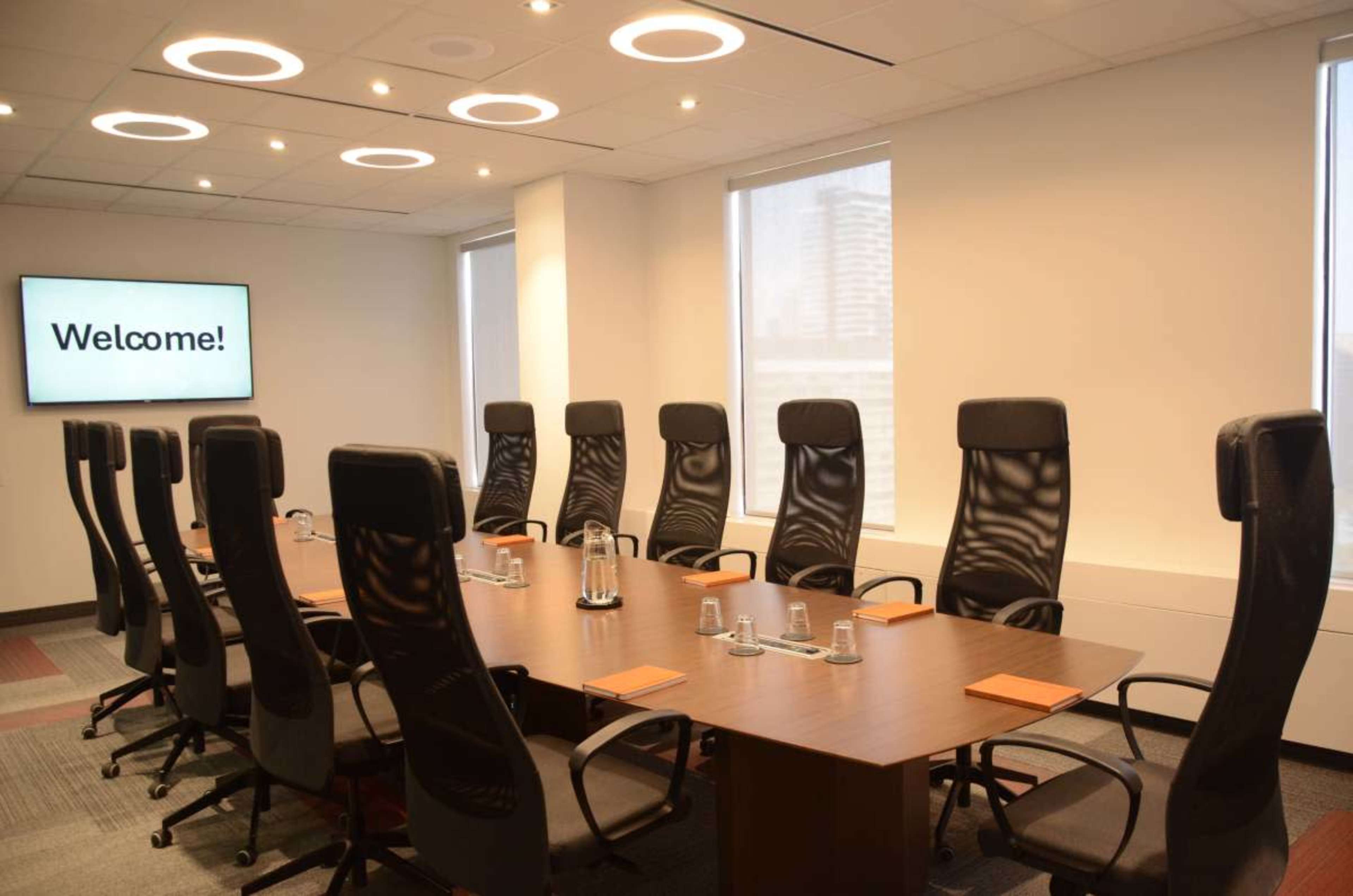 A modern conference room features a large table surrounded by ergonomic chairs, with a welcome message displayed on a screen.