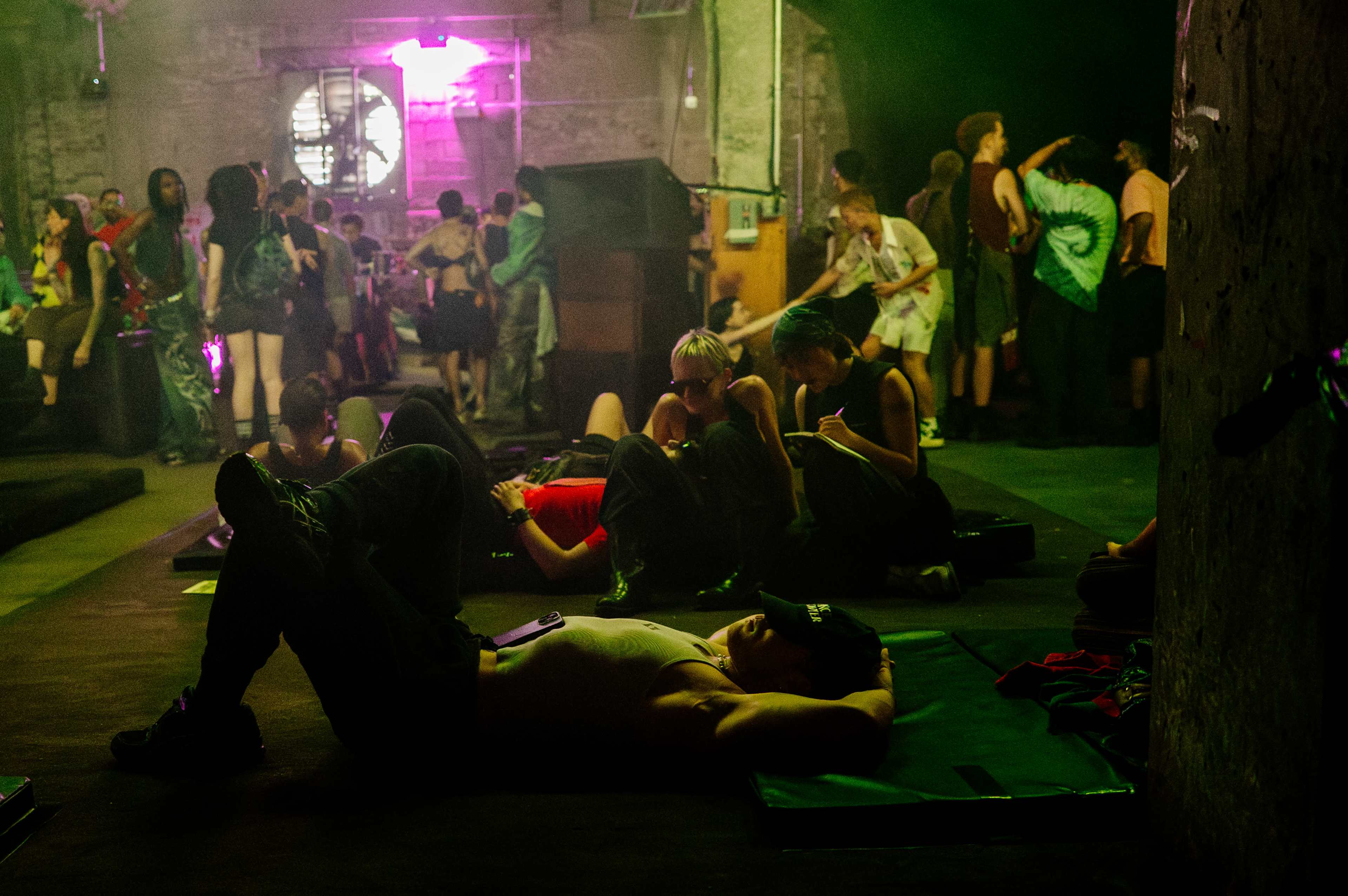 A crowded underground gathering features groups of people socializing, some lounging on the floor while others stand around in low lighting.