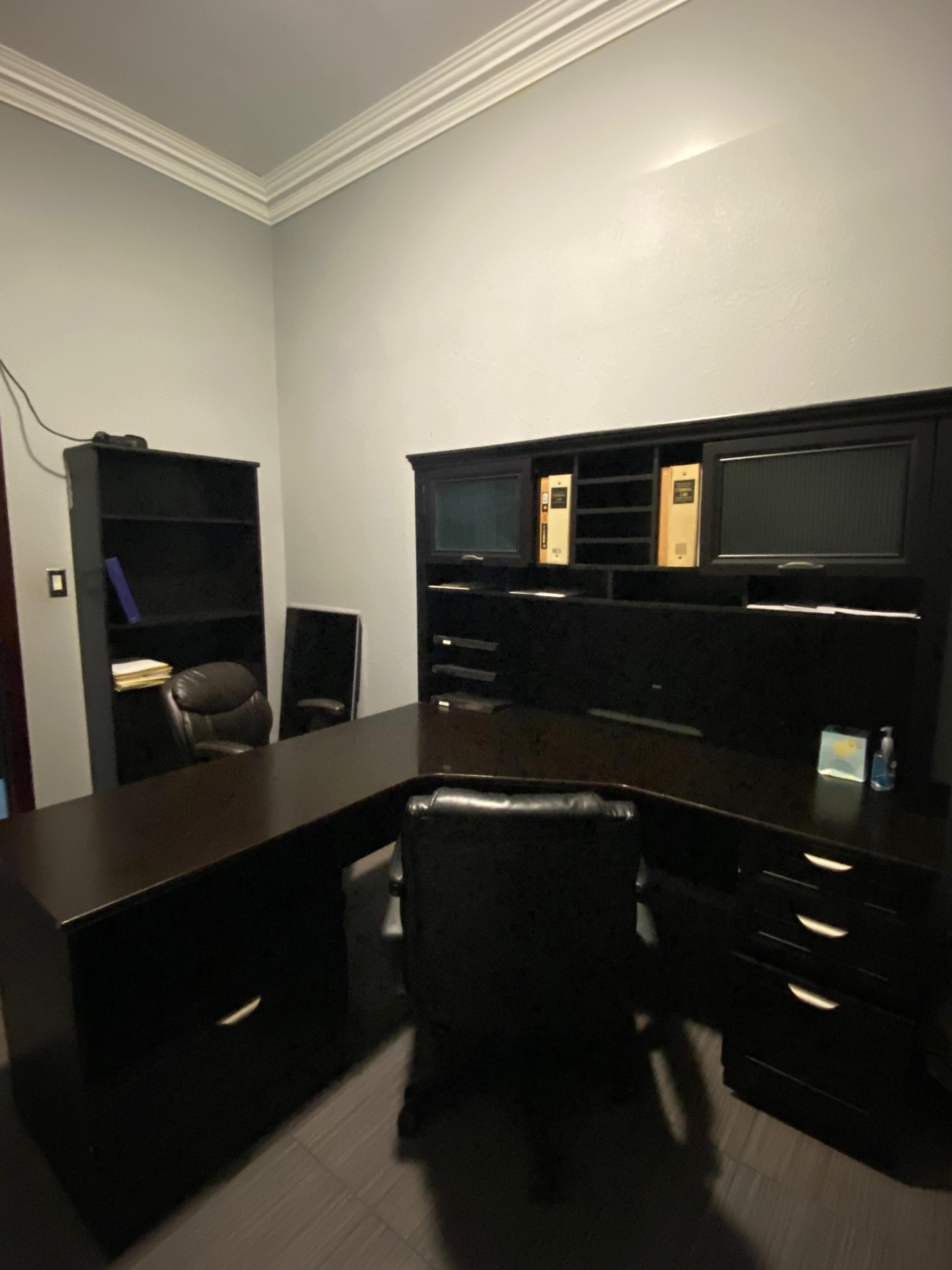 A dark wooden office setup with a large desk, an ergonomic chair, and a bookshelf in a dimly lit room.