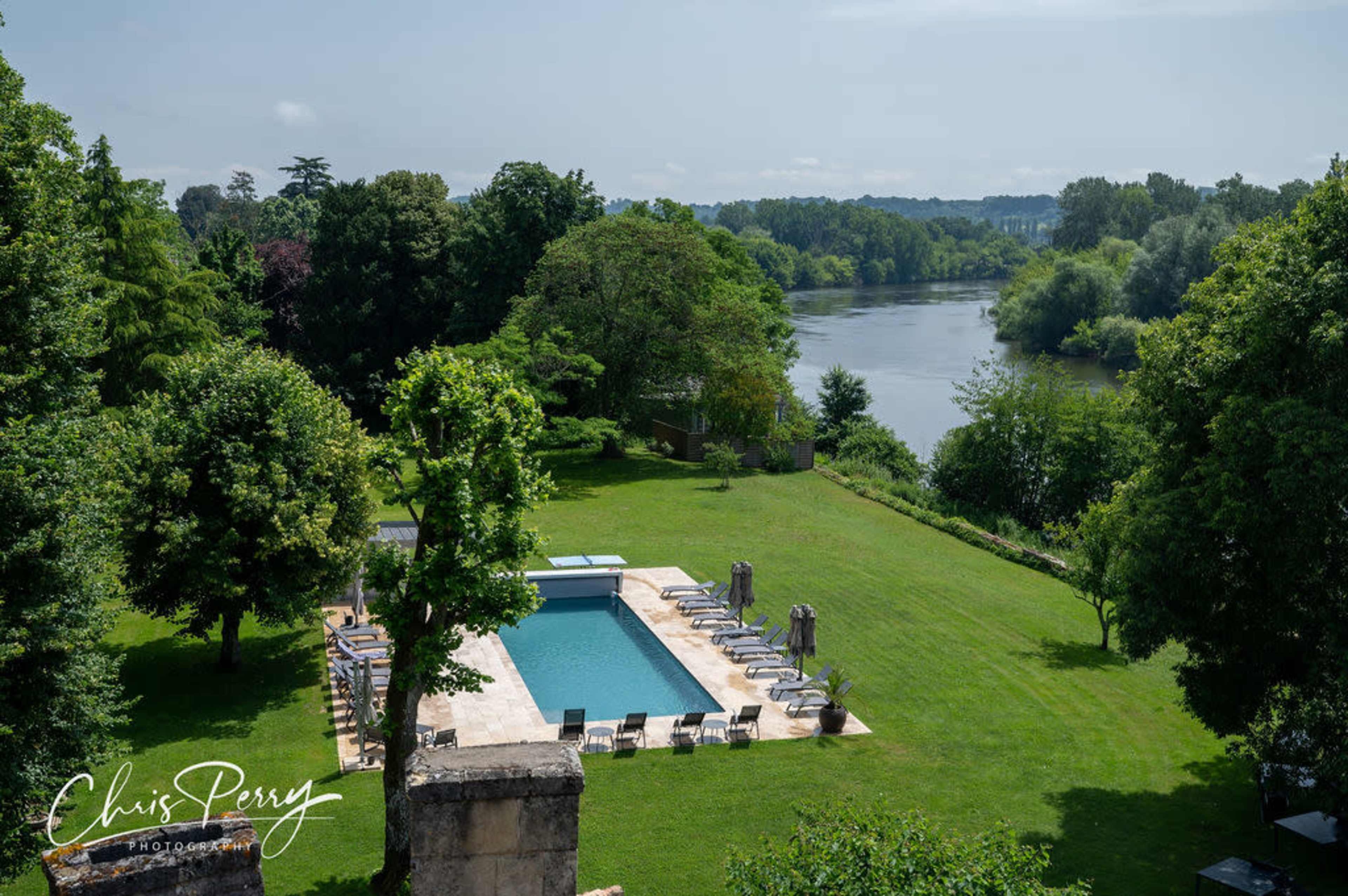 The image shows a large, green lawn with a swimming pool and lounge chairs, overlooking a river lined with trees.