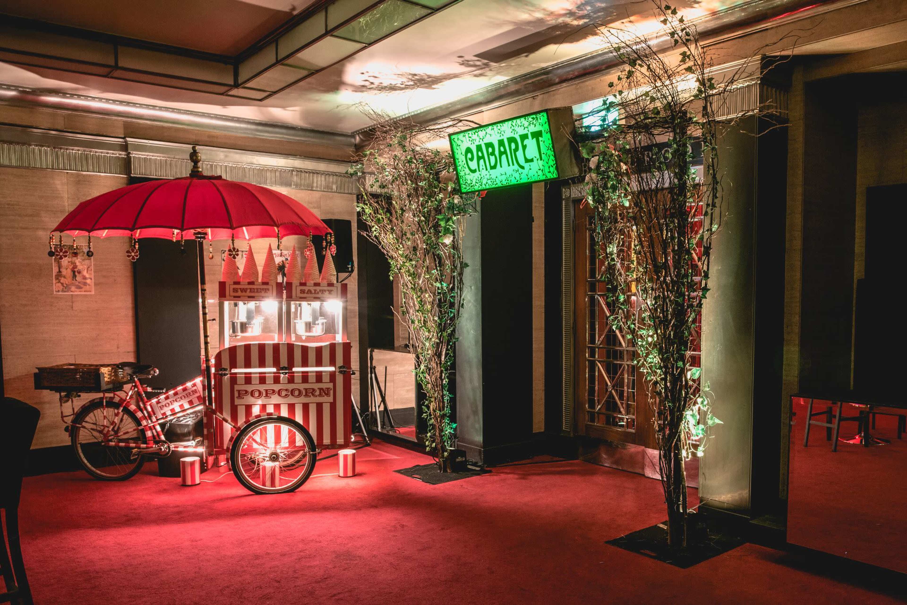 A vintage-style popcorn cart under a red umbrella, positioned on a red carpeted floor beside illuminated cabaret signage and decorative greenery.