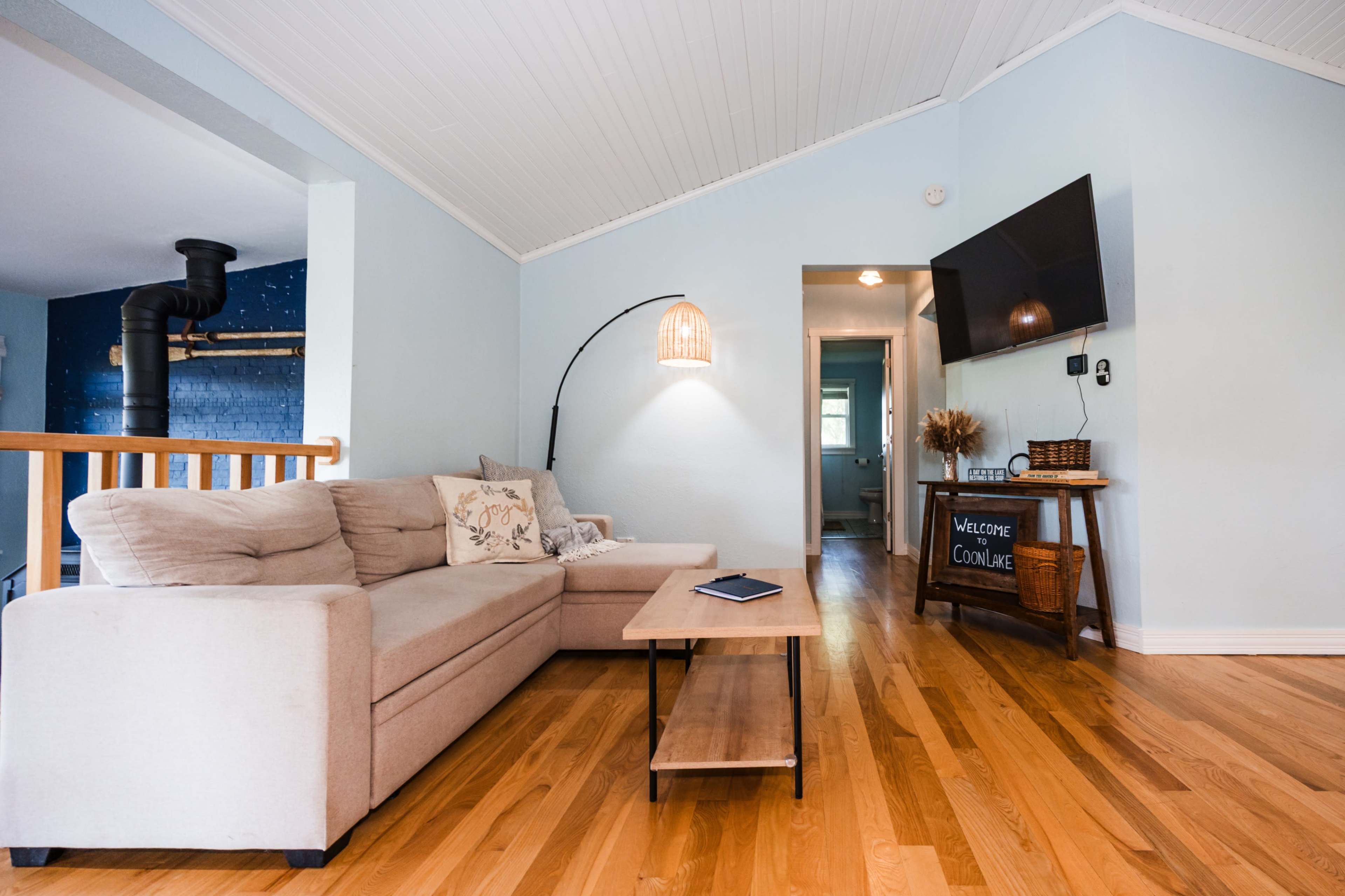 The image shows a living room with a light blue wall, a beige sectional sofa, a wooden coffee table, and a flat-screen TV mounted on the wall.