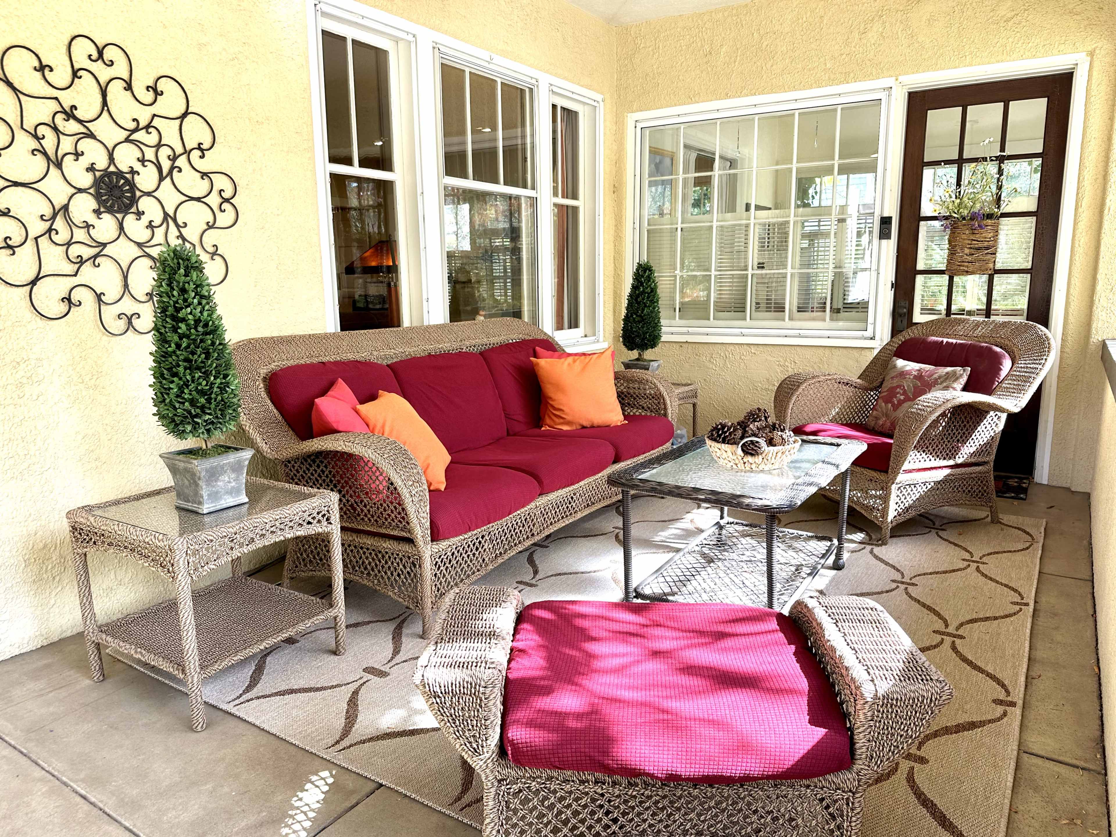 The image shows a cozy outdoor seating area with wicker furniture, colorful cushions, and a coffee table set on a patterned rug against a yellow wall.