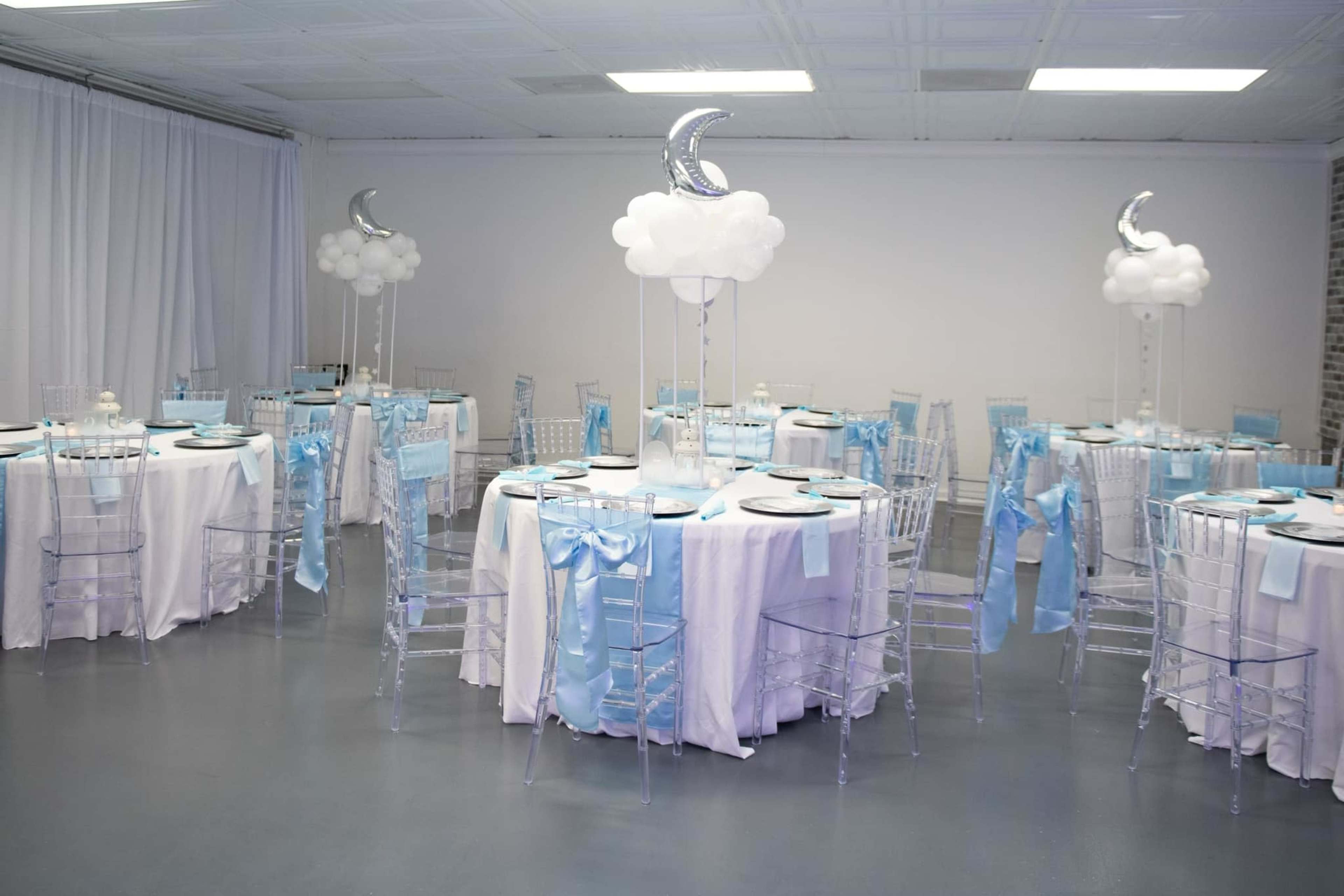 The image depicts a banquet hall set up for an event, featuring round tables covered with white linens and light blue decorative accents, along with clear chiavari chairs and balloon centerpieces resembling clouds and moons.