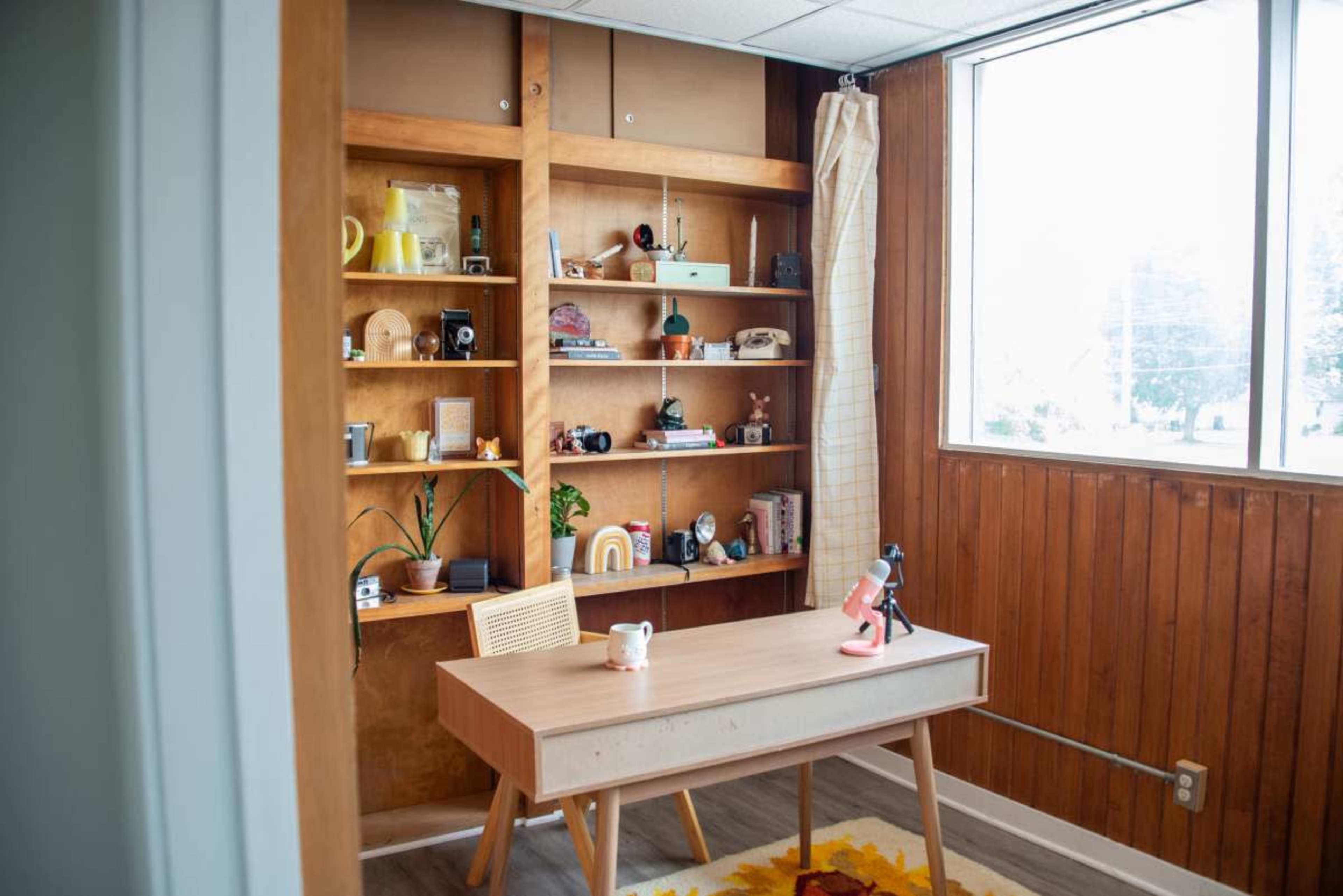 The image shows a small office with a wooden desk, a shelf displaying various decorative items, and a window letting in natural light.