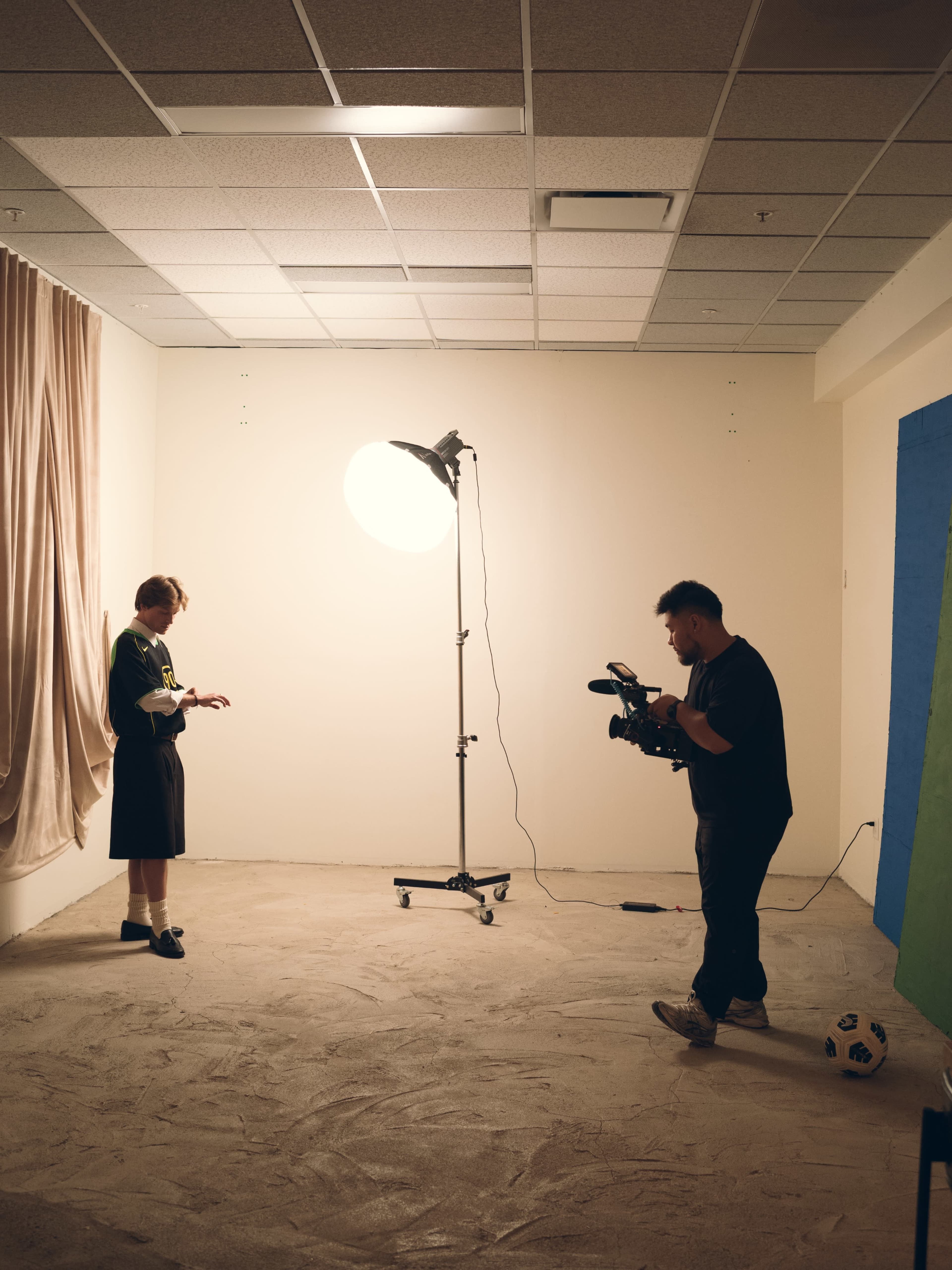 A photographer operates a camera while a subject stands by a light in a sparse studio setting with beige walls and a sandy floor.
