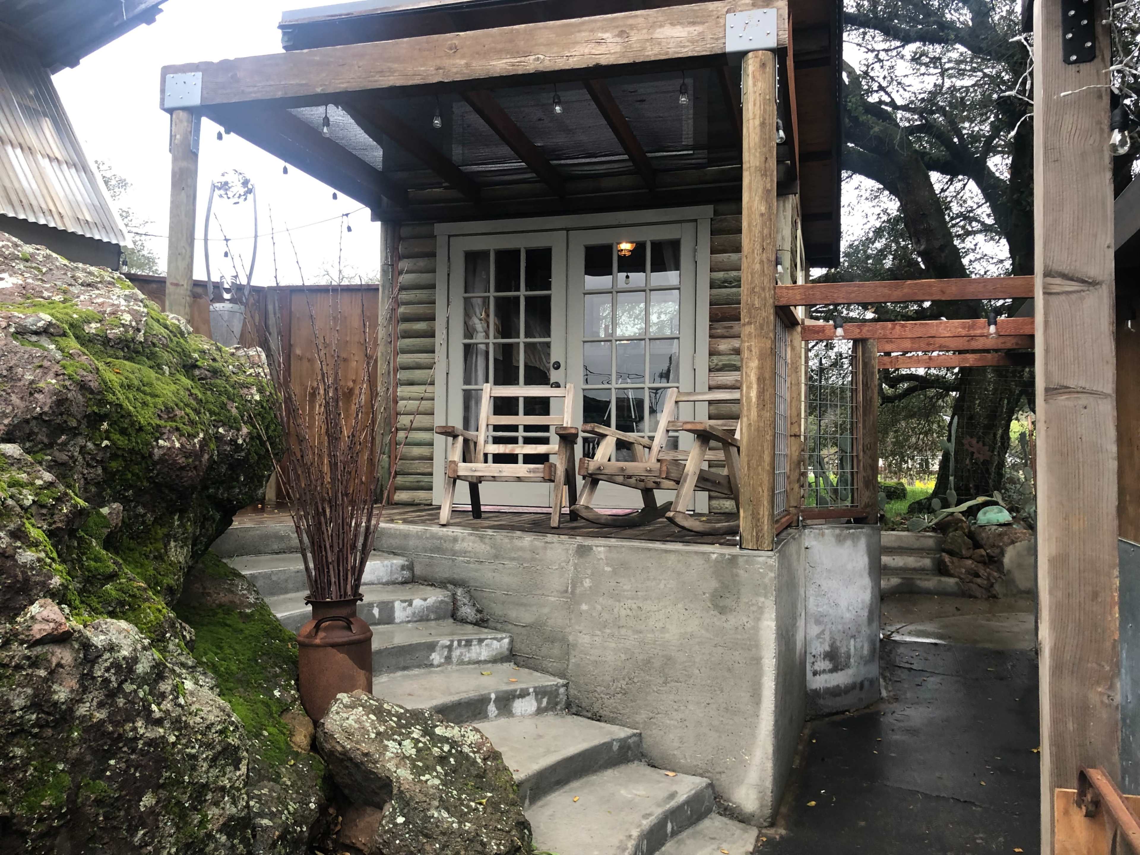 A small wooden cabin with a covered porch and two chairs, surrounded by large rocks and greenery.