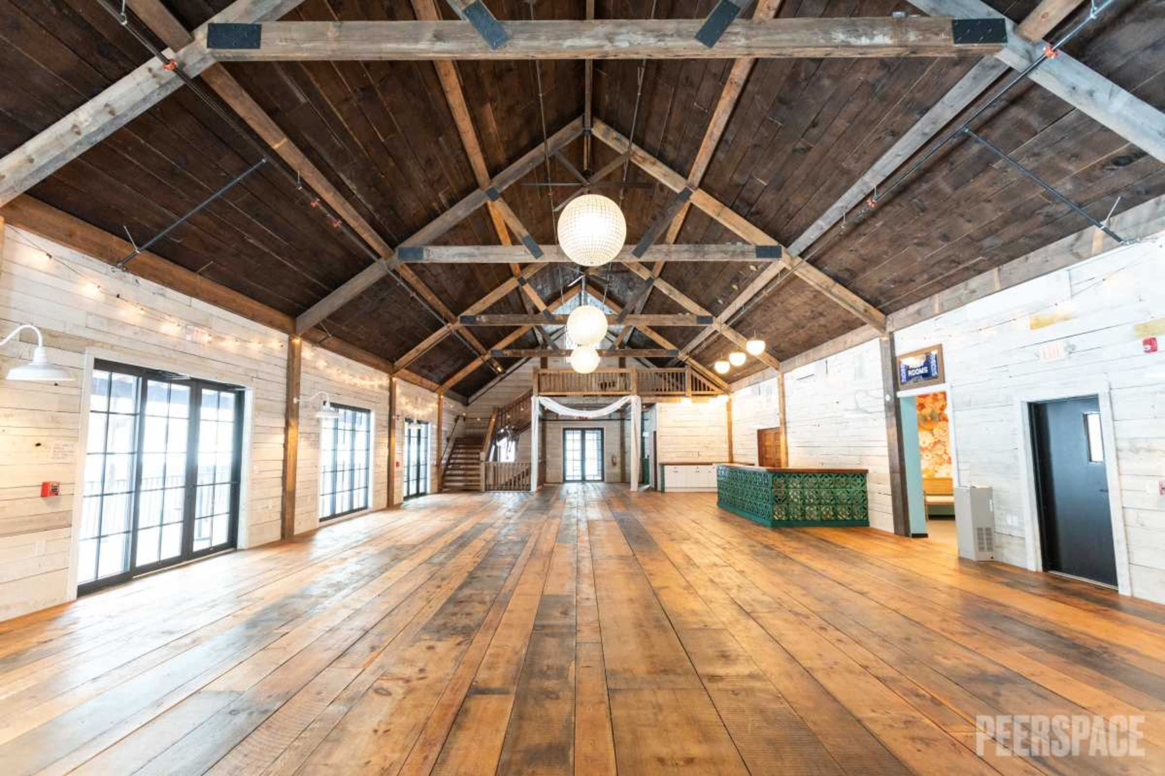 The image shows a spacious, wooden interior of a hall with high ceilings, large windows, and a green reception desk towards the back.