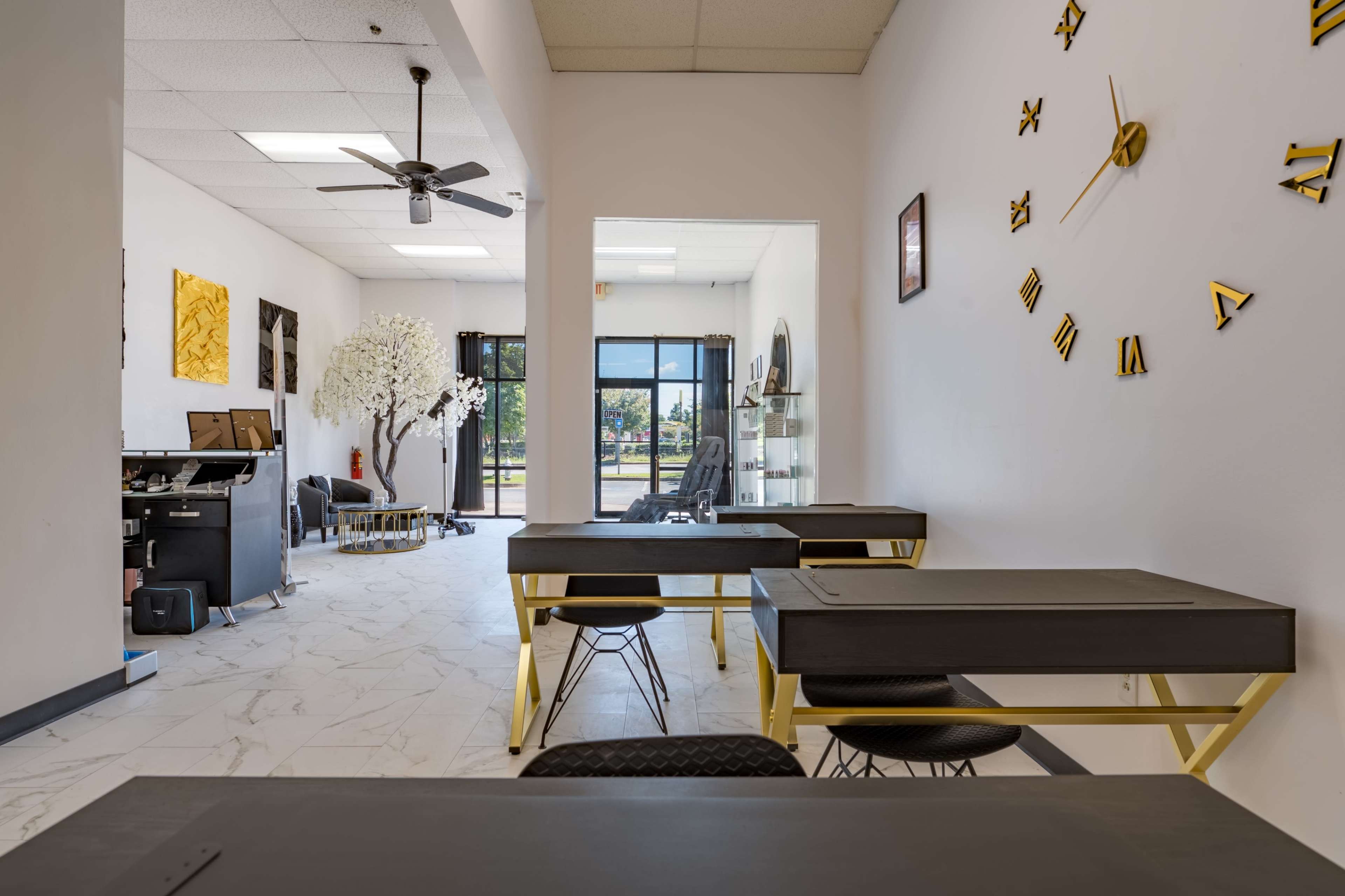 A modern workspace featuring several gray desks with gold legs, a large open area, and a decorative tree, with glass doors leading to an outdoor view.