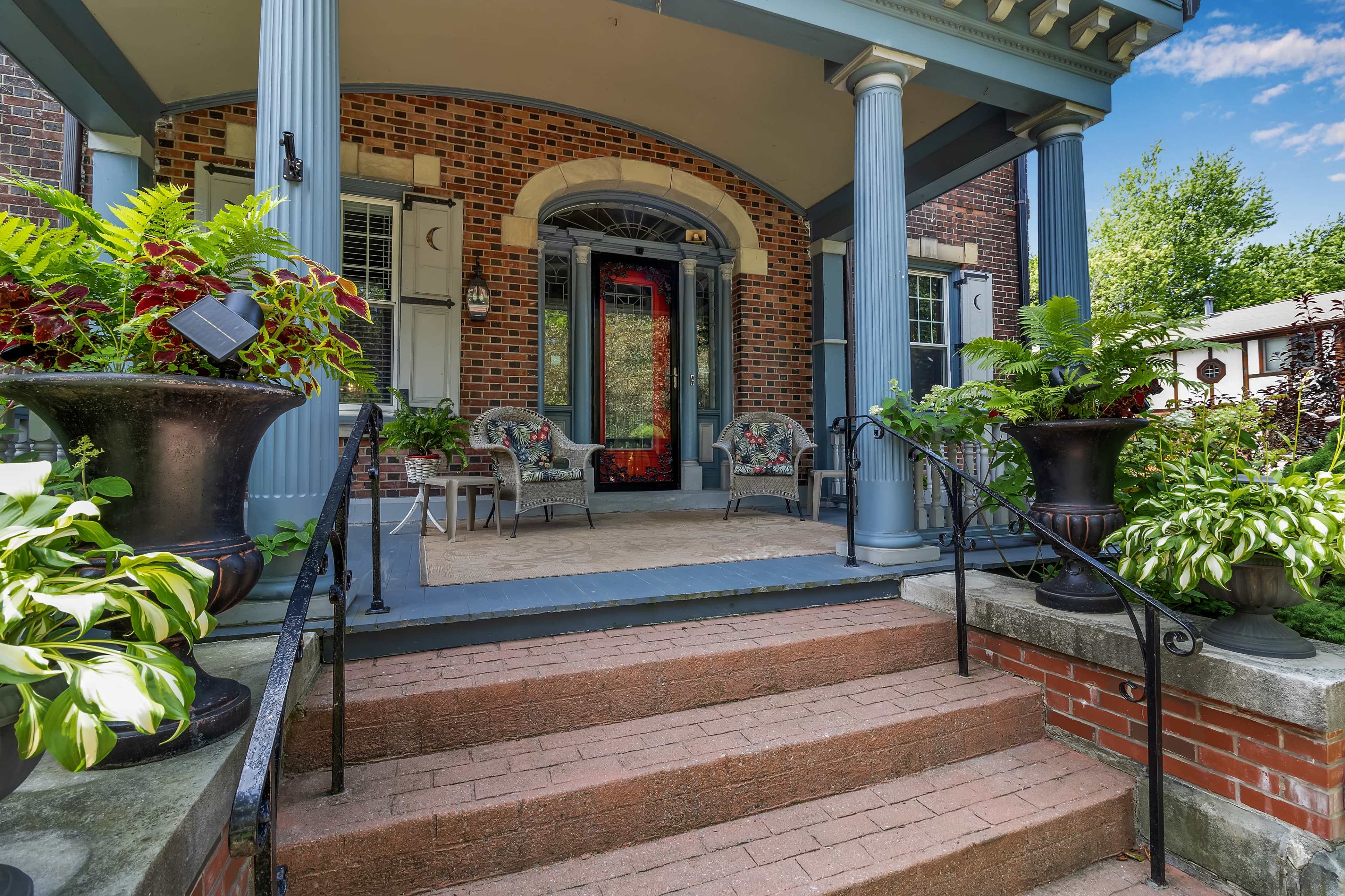 A brick porch with two decorative chairs and planters filled with greenery beside a set of stairs.