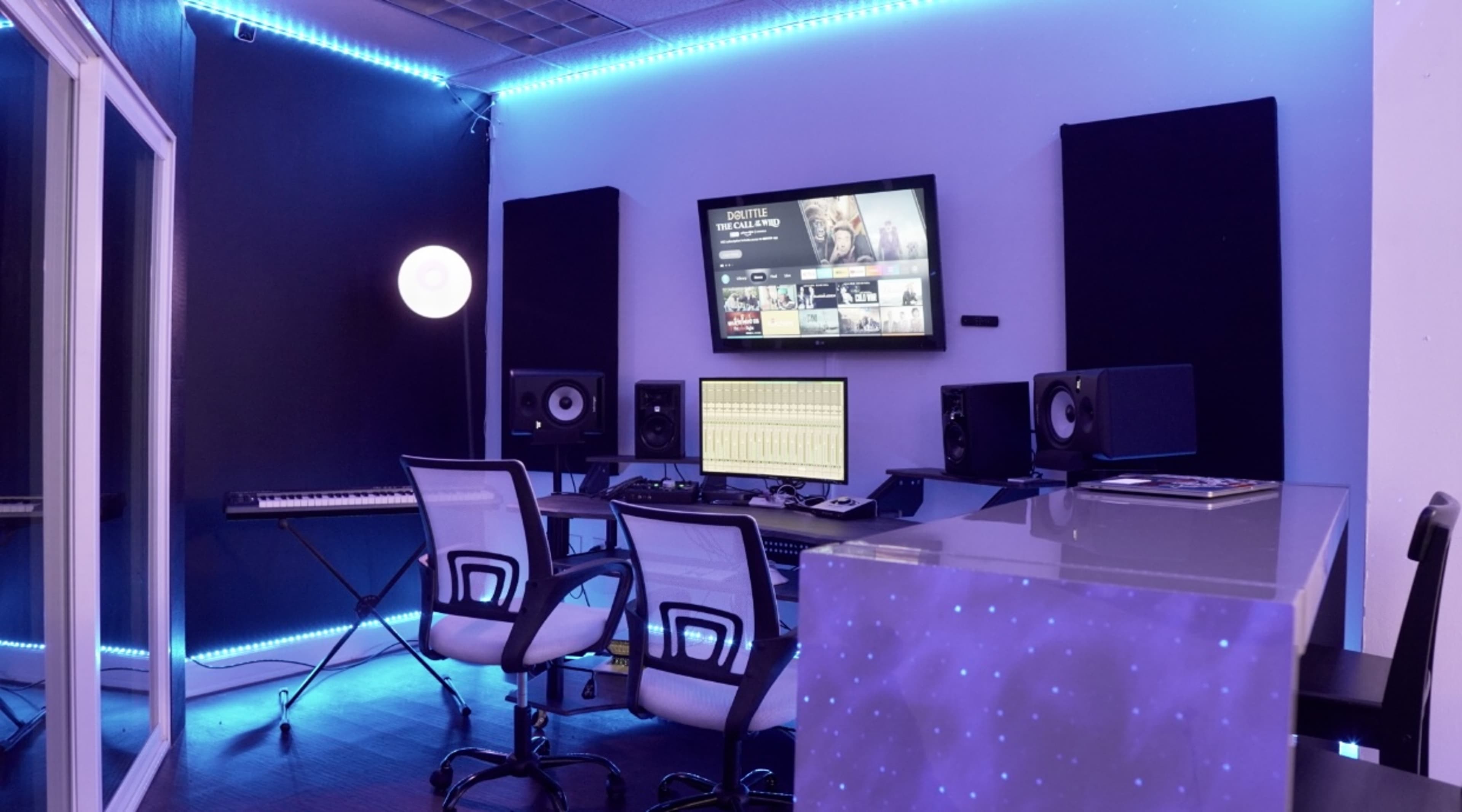 The image shows a modern music production studio featuring a desk with two chairs, studio monitors, a keyboard, a large screen mounted on the wall, and ambient LED lighting.