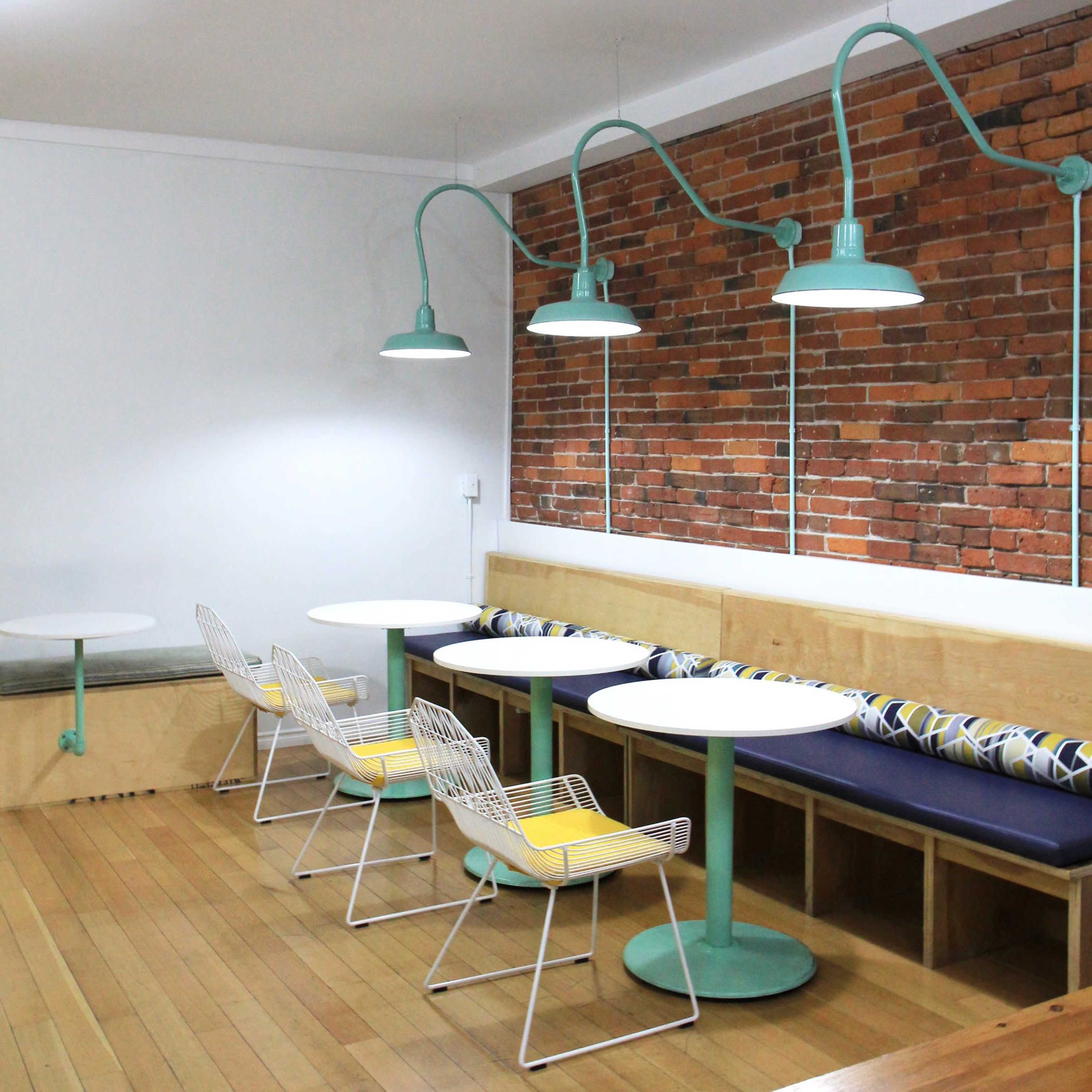 A modern café interior with two round tables, wire chairs with yellow cushions, and pendant lights hanging from the ceiling above a brick wall.