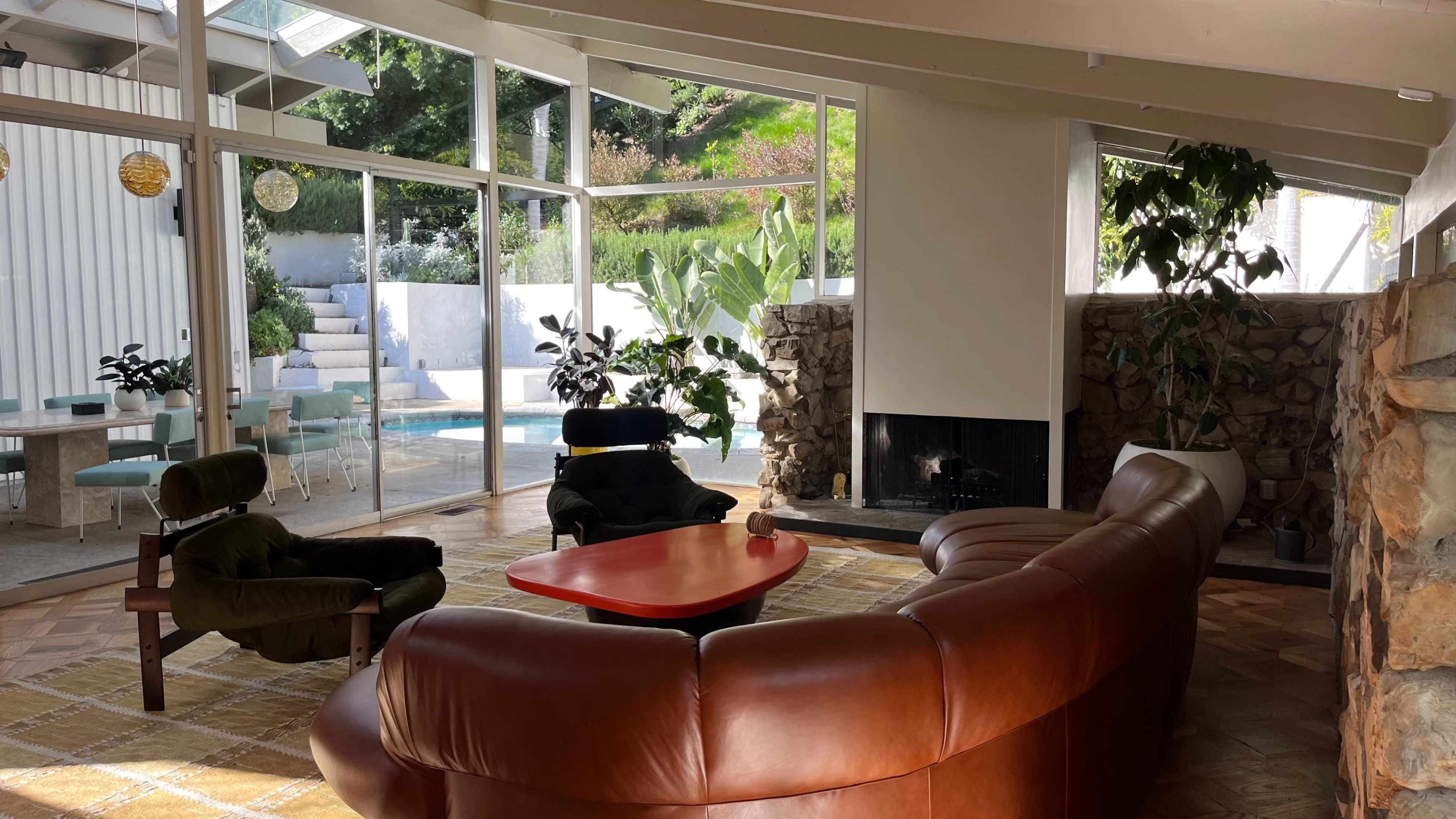 A spacious living room features a brown leather sectional sofa, a red coffee table, and large windows overlooking a pool area surrounded by greenery.