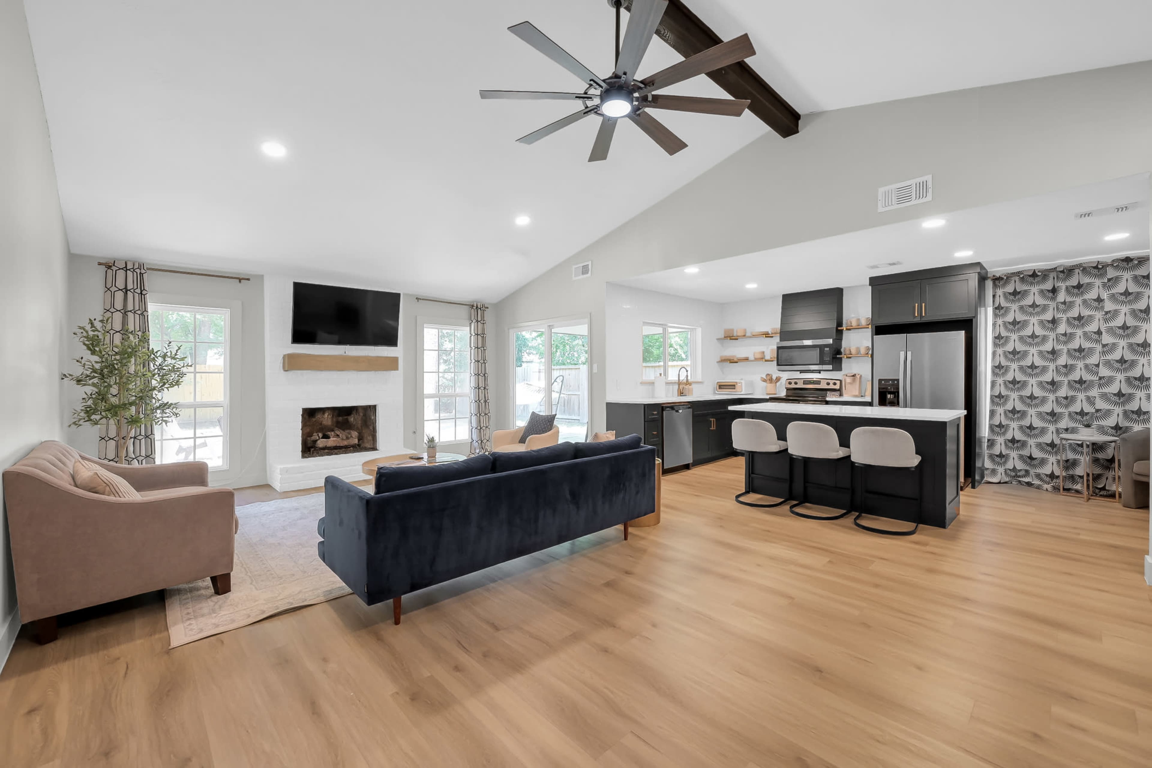 The image shows a modern living room with a fireplace, a large window, and an open kitchen area featuring a dark island and bar stools.