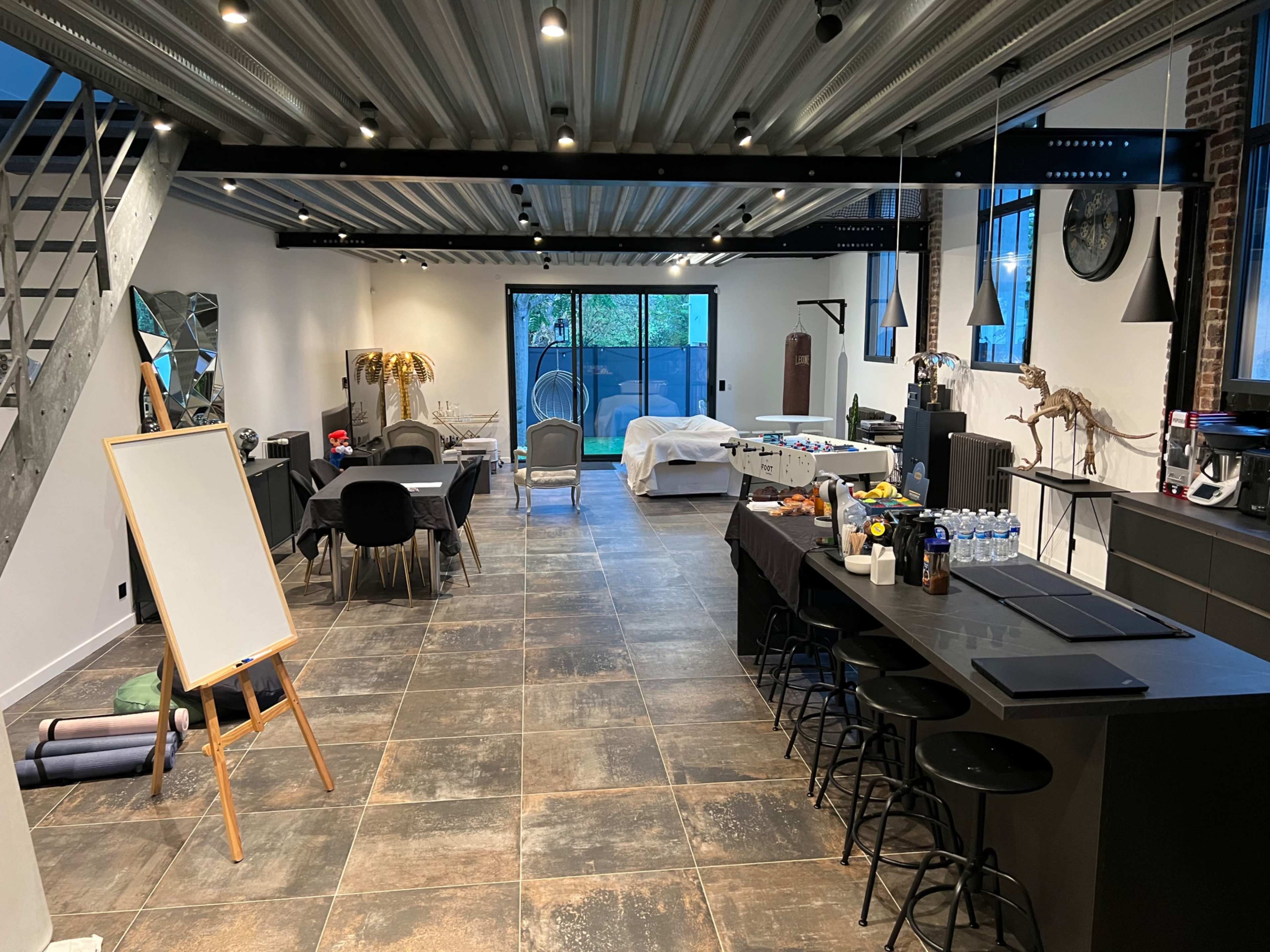 The image shows a spacious, modern room with a kitchen area, dining space, and various furnishings, illuminated by overhead lights and natural light from large windows.