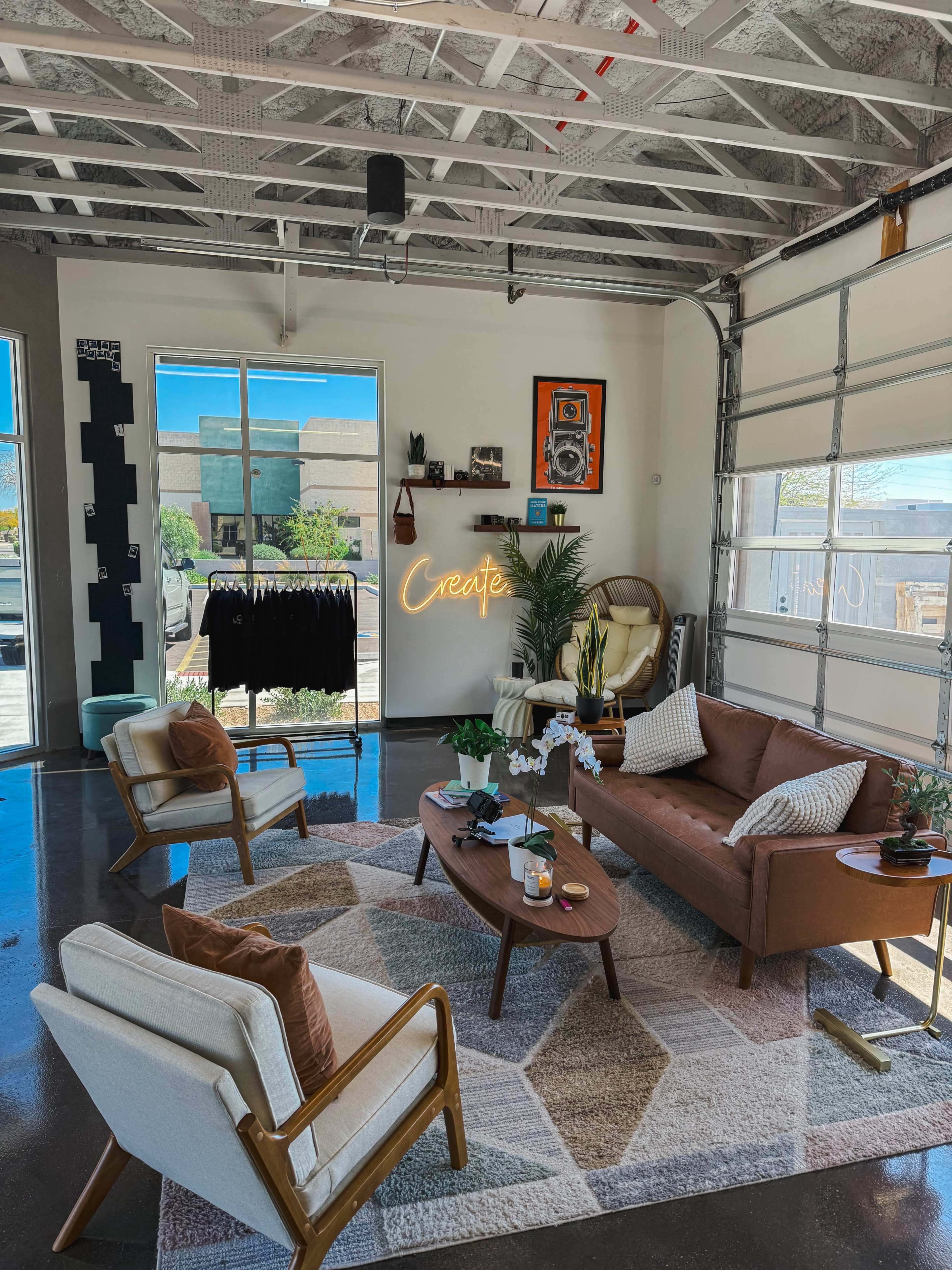 The image shows a modern living room with a mix of furniture, including a brown sofa, two light-colored armchairs, and a coffee table, along with a "Create" neon sign on the wall and a clothing rack nearby.