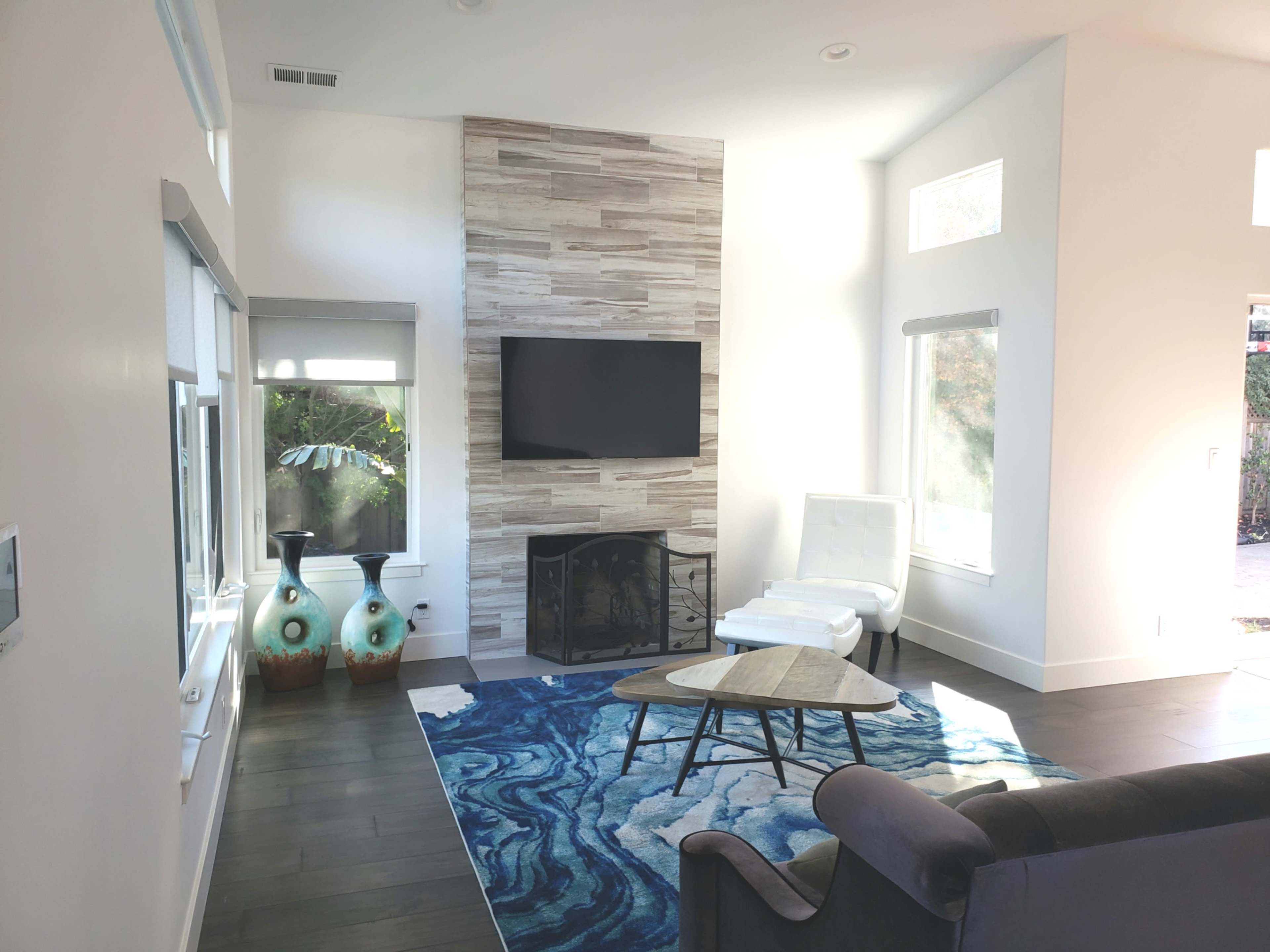 A modern living room features a stone accent wall with a mounted TV, a white chair, a round coffee table on a blue rug, and two decorative vases.