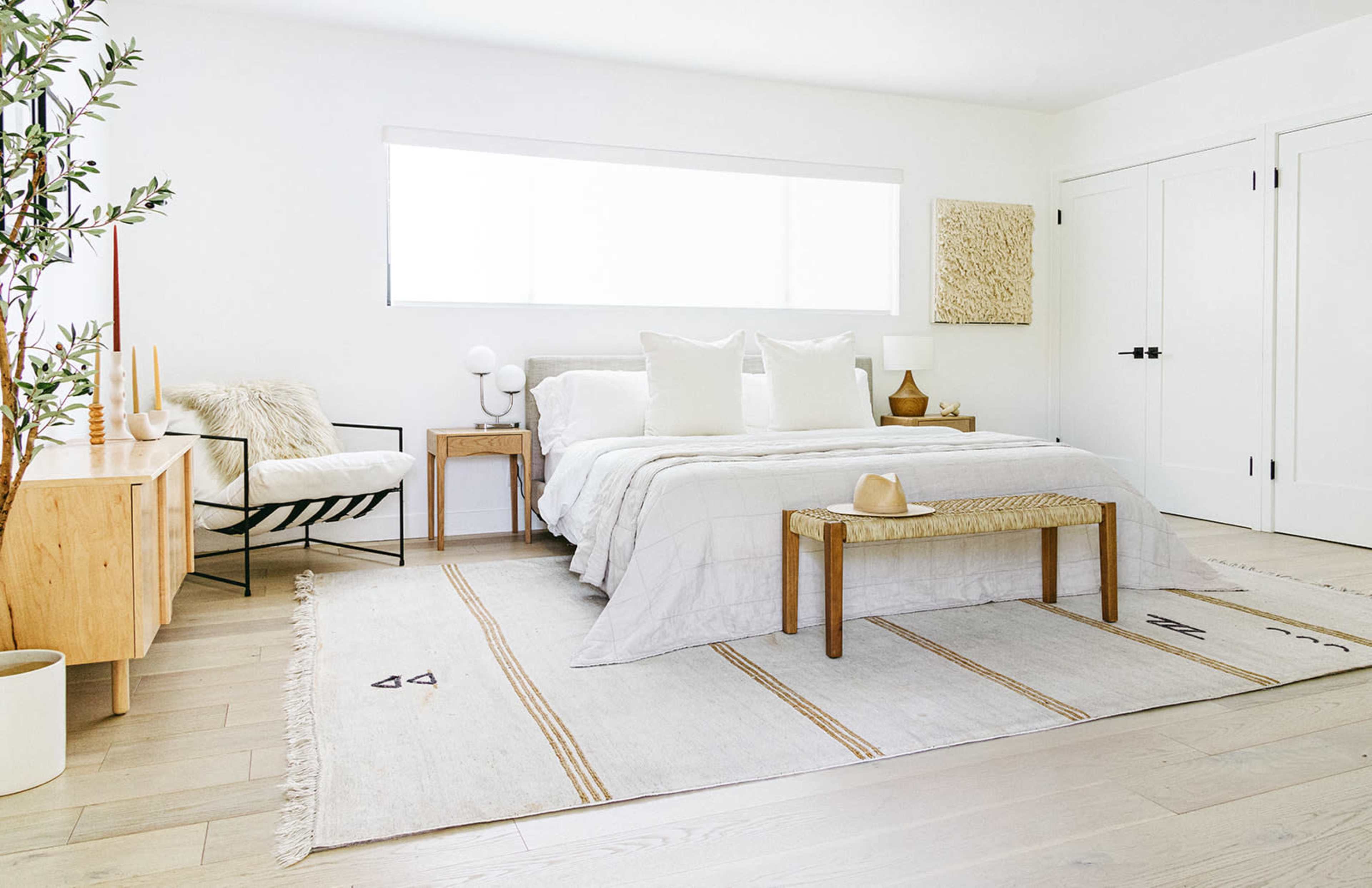 A neatly arranged modern bedroom features a bed with white linens, a wooden bench at the foot, and minimalist furniture against bright walls.