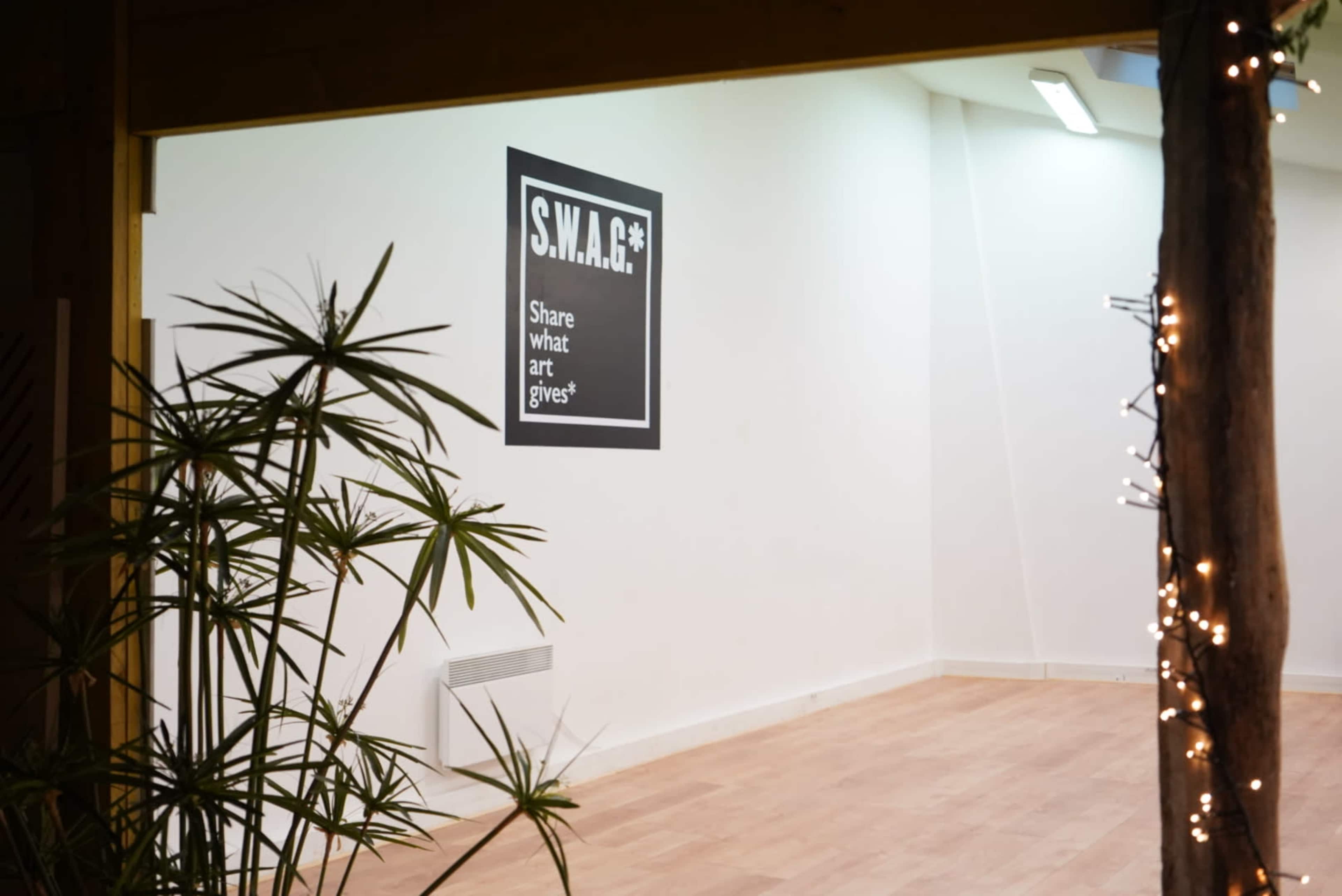 A minimalist interior space featuring a large wall sign that reads "S.W.A.G.: Share What Art Gives," with potted plants in the foreground.