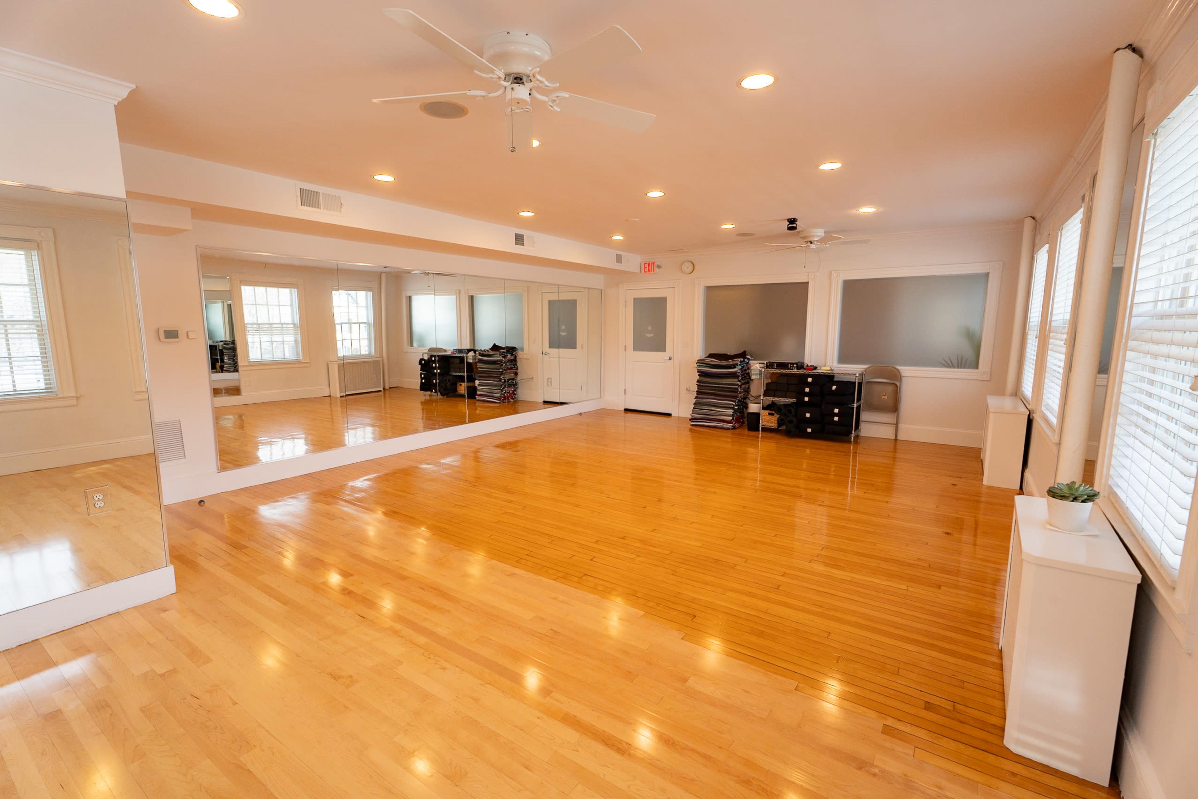 The image shows a spacious room with hardwood floors, mirrors on one wall, and several ceiling lights, creating a bright and open environment.