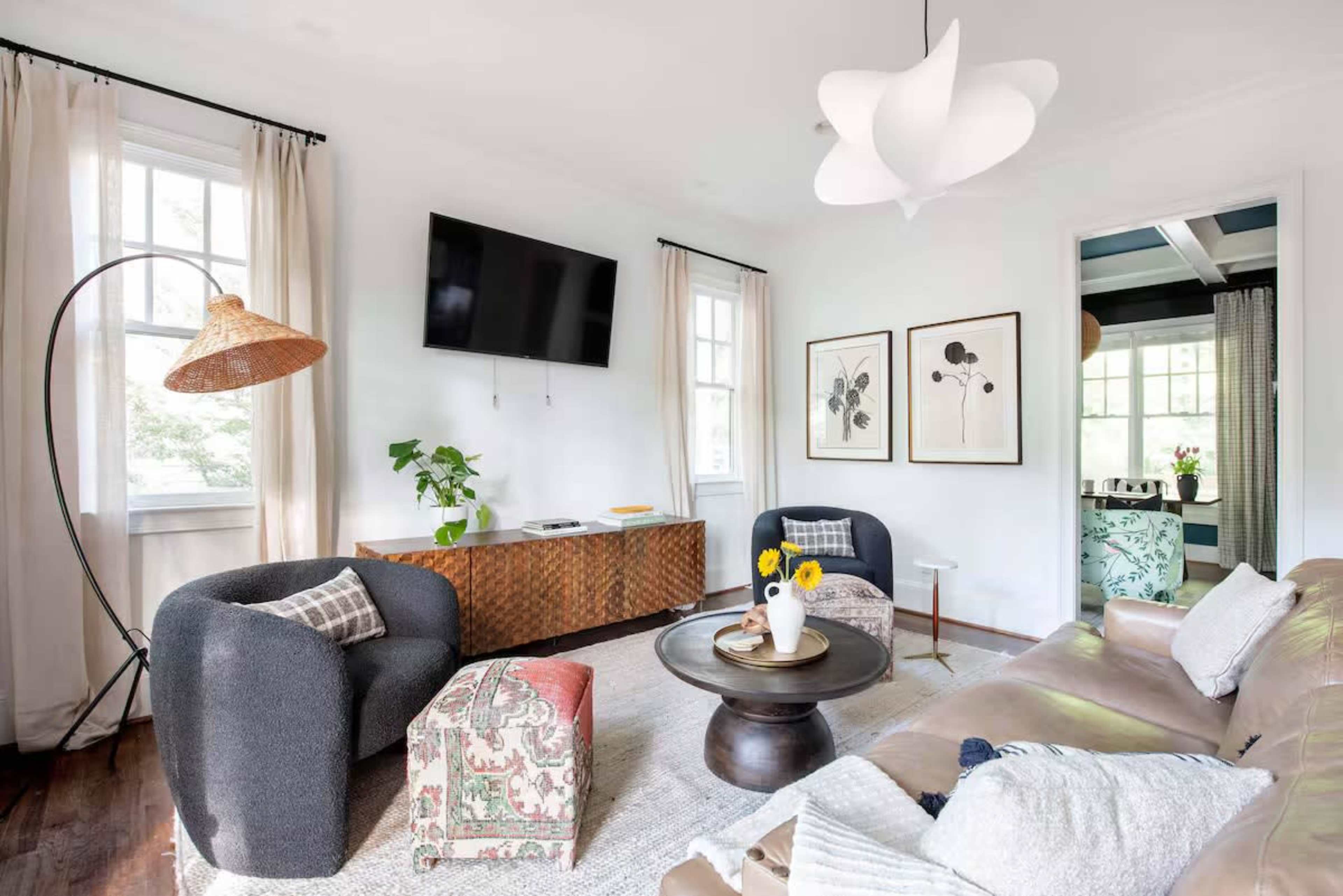 The image shows a modern living room with a light-colored couch, a round coffee table, two armchairs, and a television on the wall, complemented by natural light from large windows.