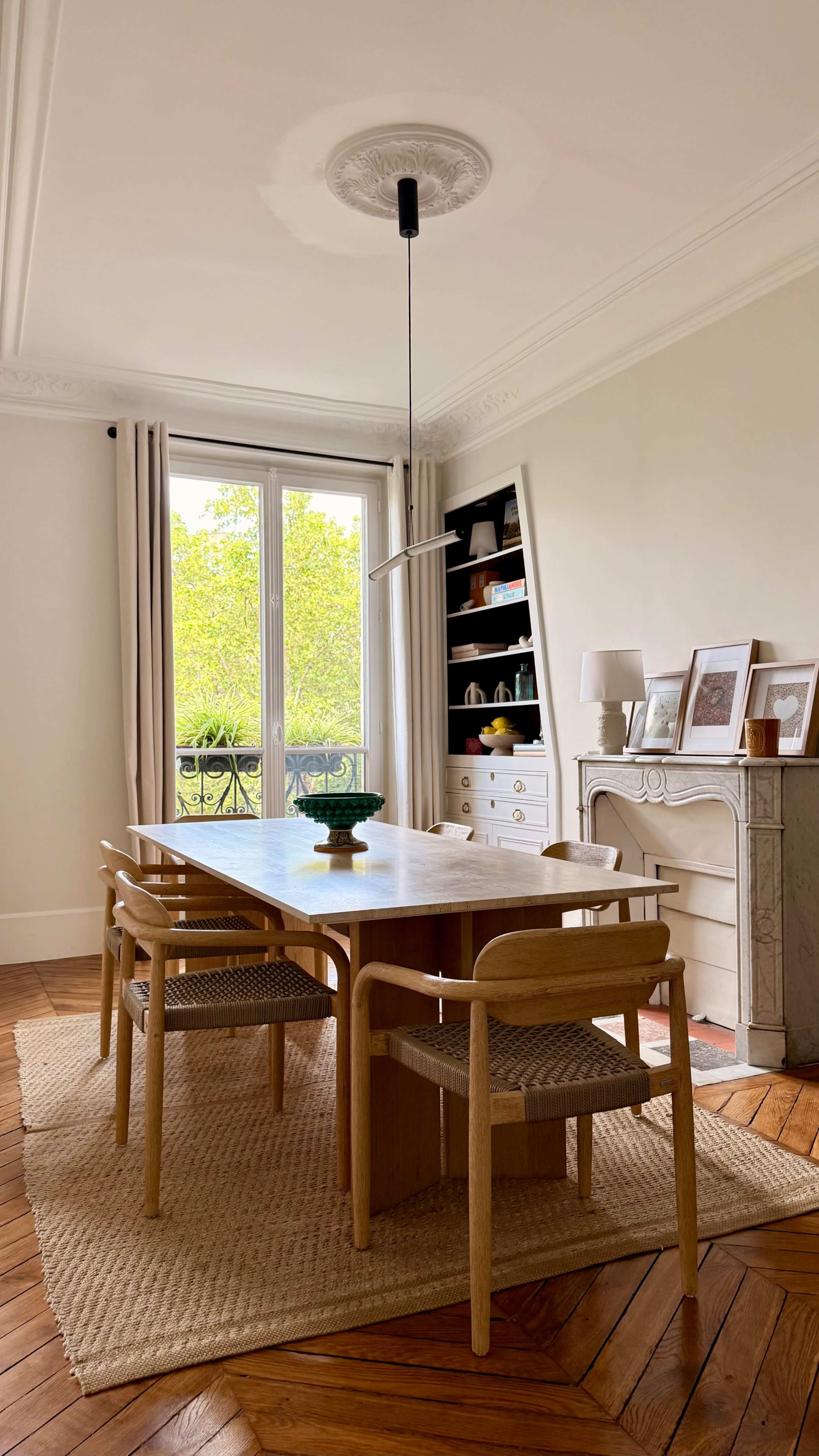 A dining room features a wooden table surrounded by four chairs, with a window providing light and a view of greenery.