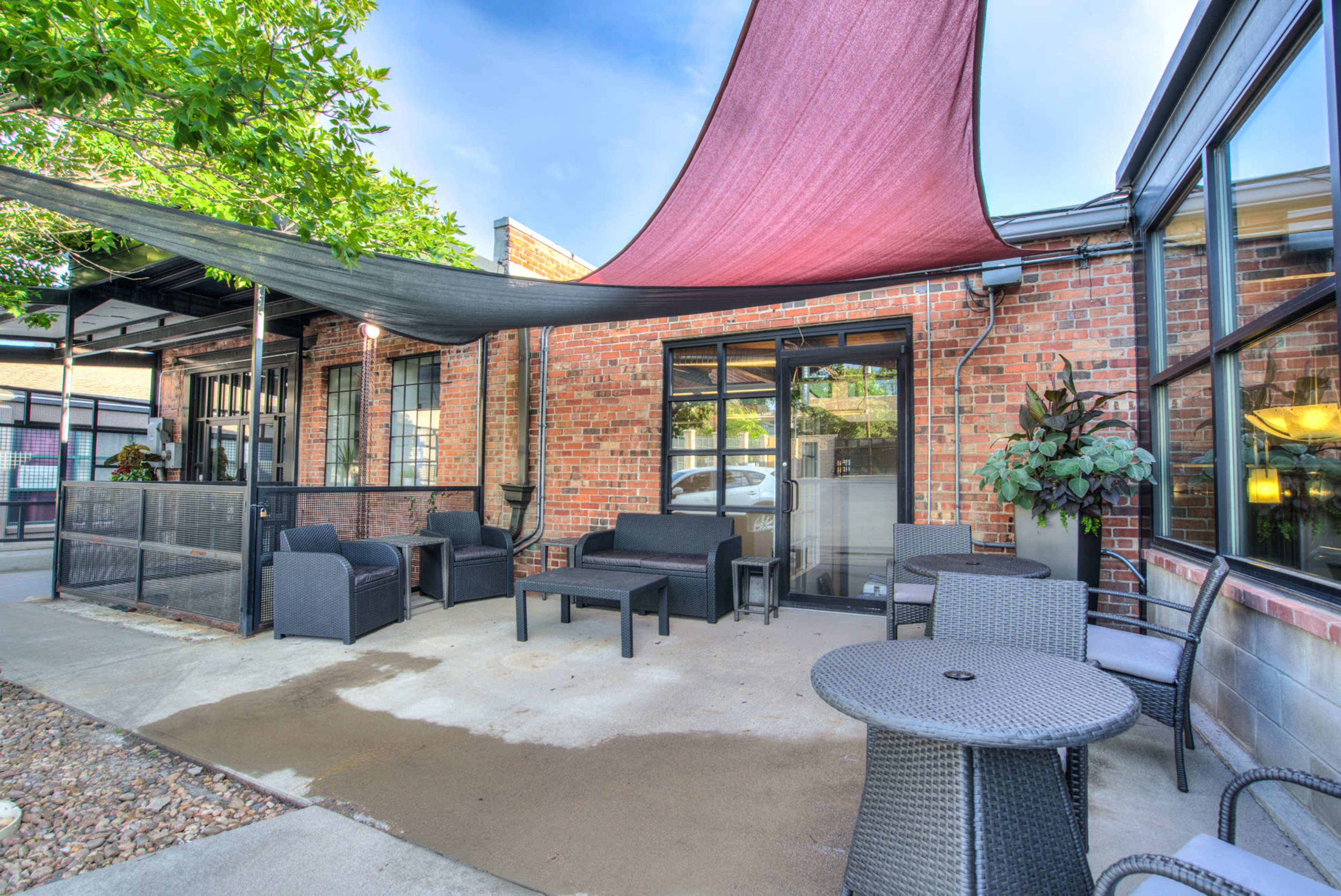 A patio with outdoor furniture, including chairs and tables, covered by colorful fabric canopies and surrounded by brick walls and potted plants.