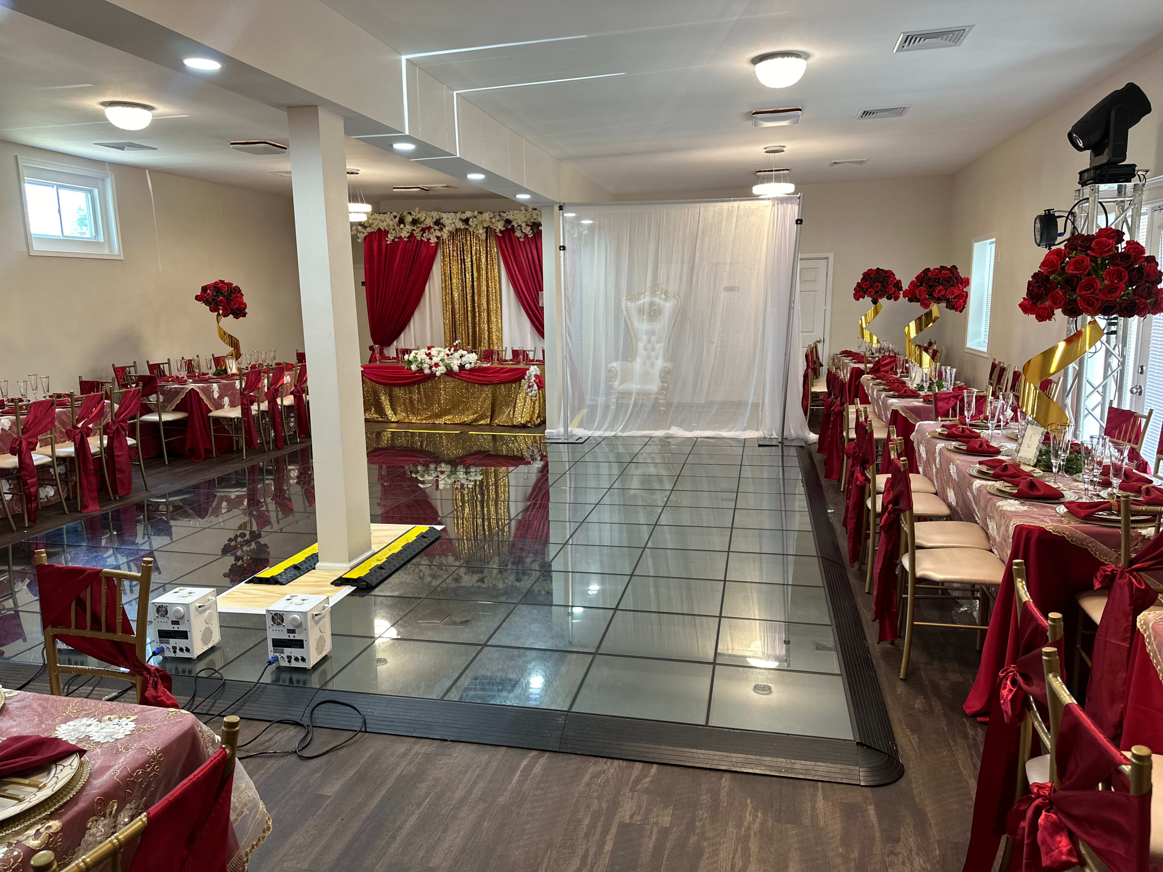 A banquet hall decorated with red and gold elements, featuring tables set with tablecloths and floral arrangements, and a dance floor illuminated in front of a backdrop with curtains.