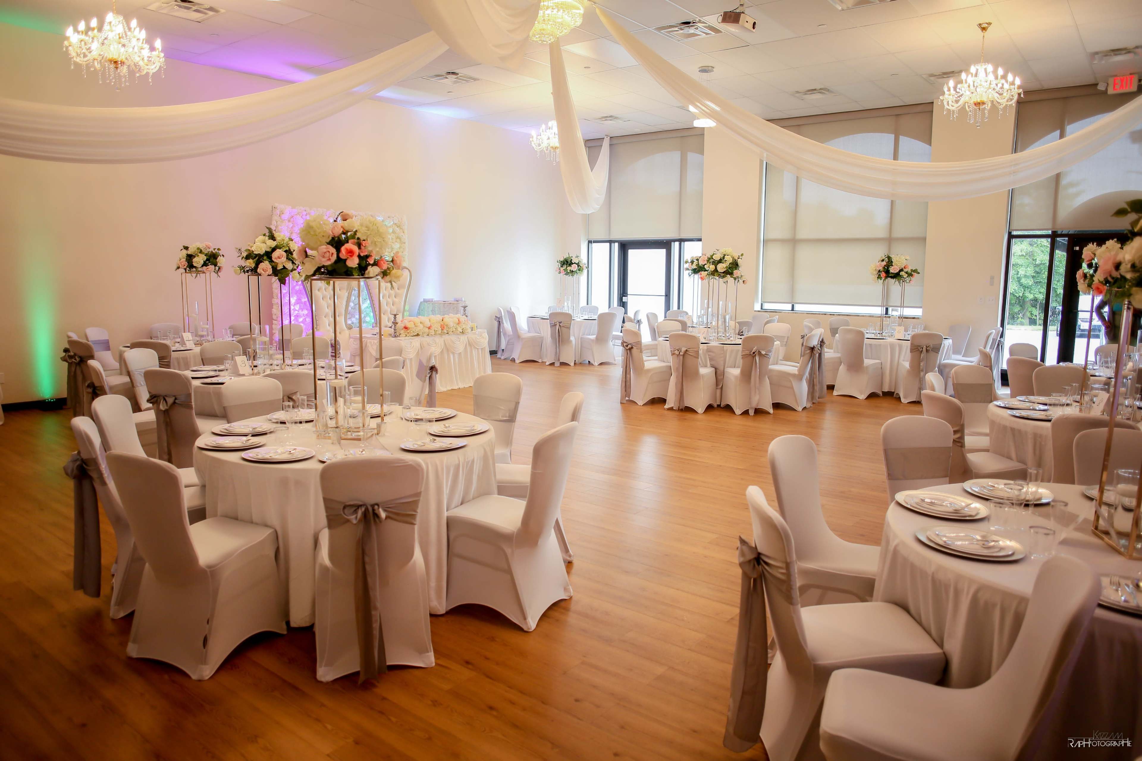 The image shows a spacious banquet hall set up for an event, featuring round tables covered with white tablecloths, elegantly arranged with centerpieces, and decorated with draped fabric and chandeliers.
