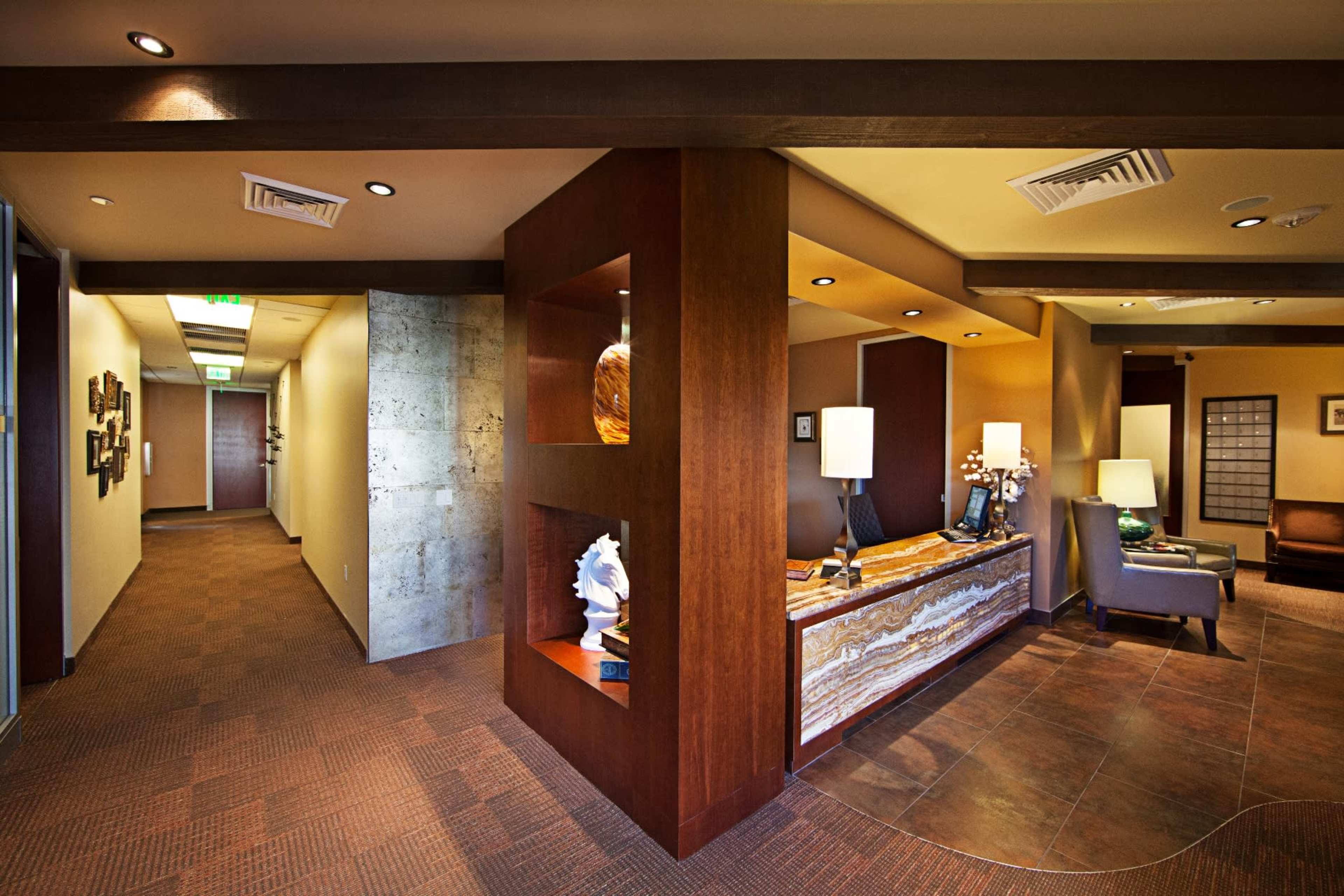 A modern office reception area featuring a wooden reception desk with lamps and a textured wall, with a corridor leading to additional office spaces.