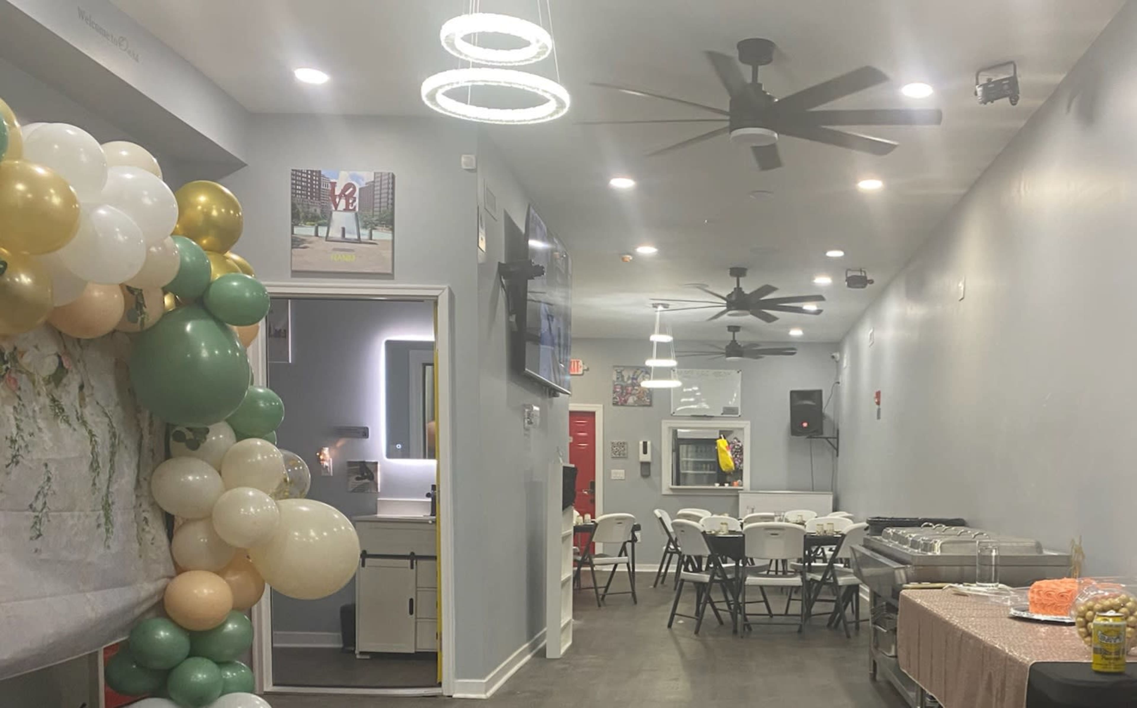 The image shows a decorated indoor event space with balloons in various colors, tables with chairs, and a kitchen area visible in the background.