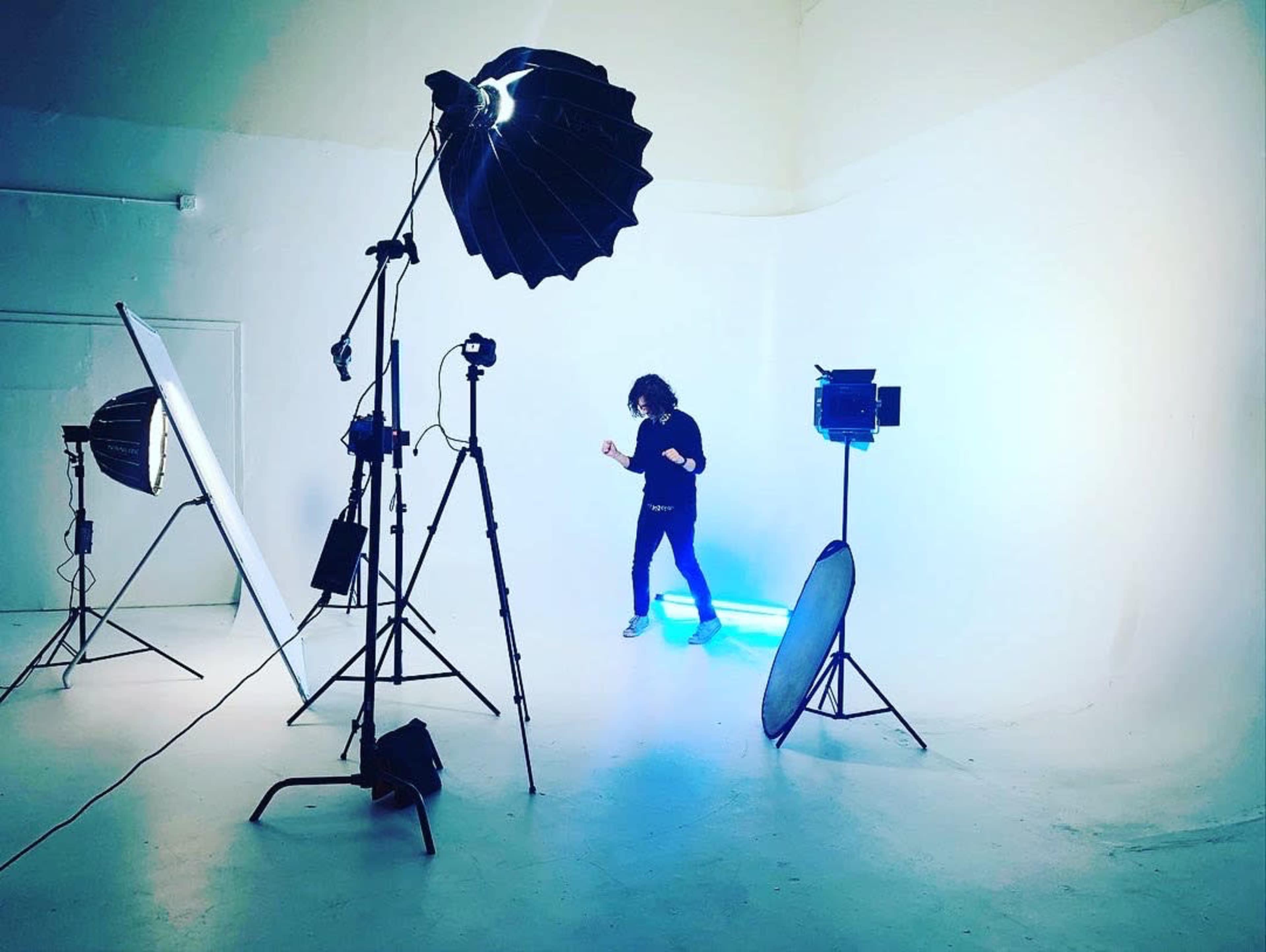 A person is dancing in a well-lit photo studio surrounded by various lighting equipment on a white backdrop.