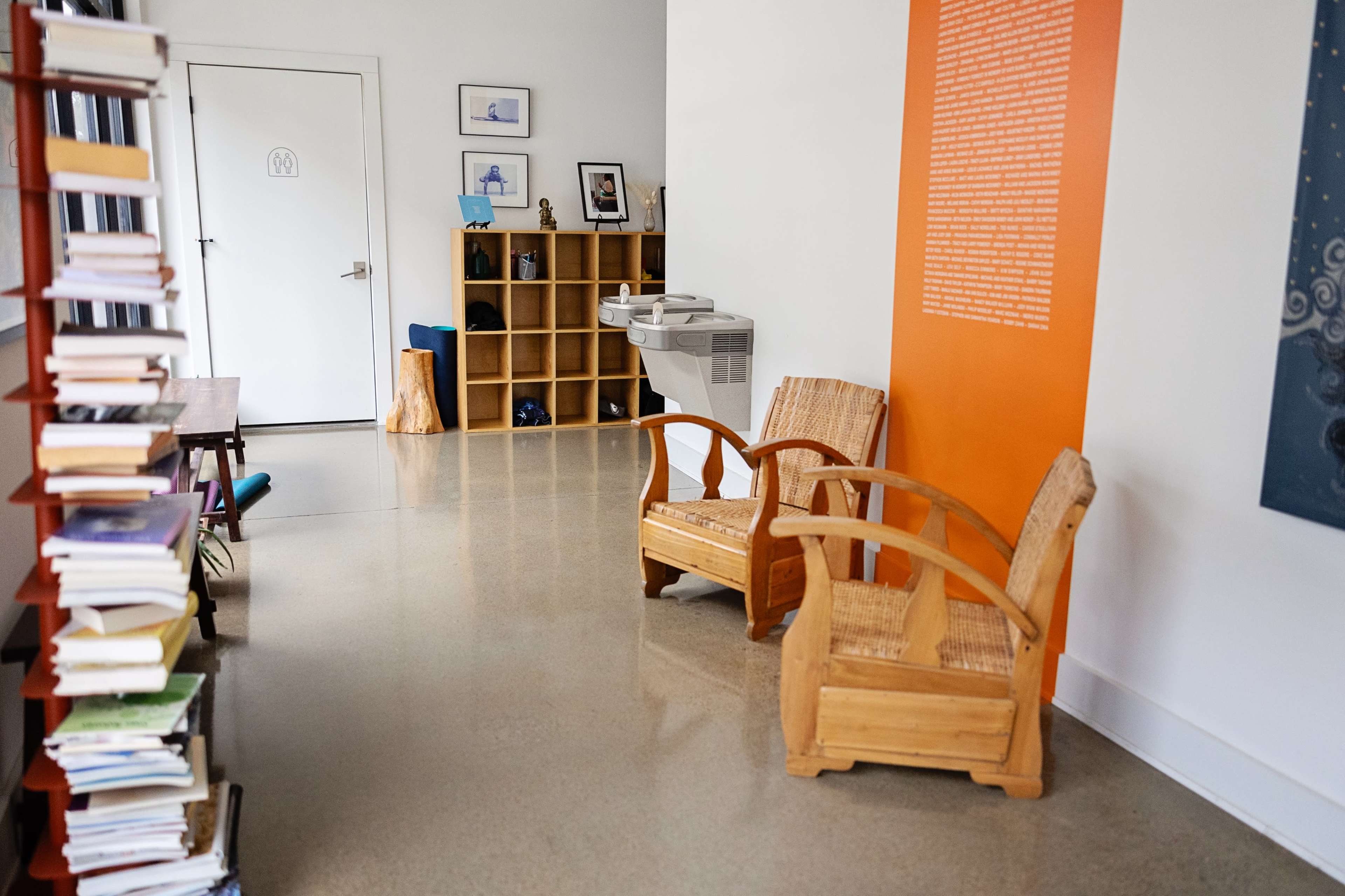 The image shows a modern interior space with two wooden chairs, a bookshelf filled with books, a water fountain, and a door leading to another room.