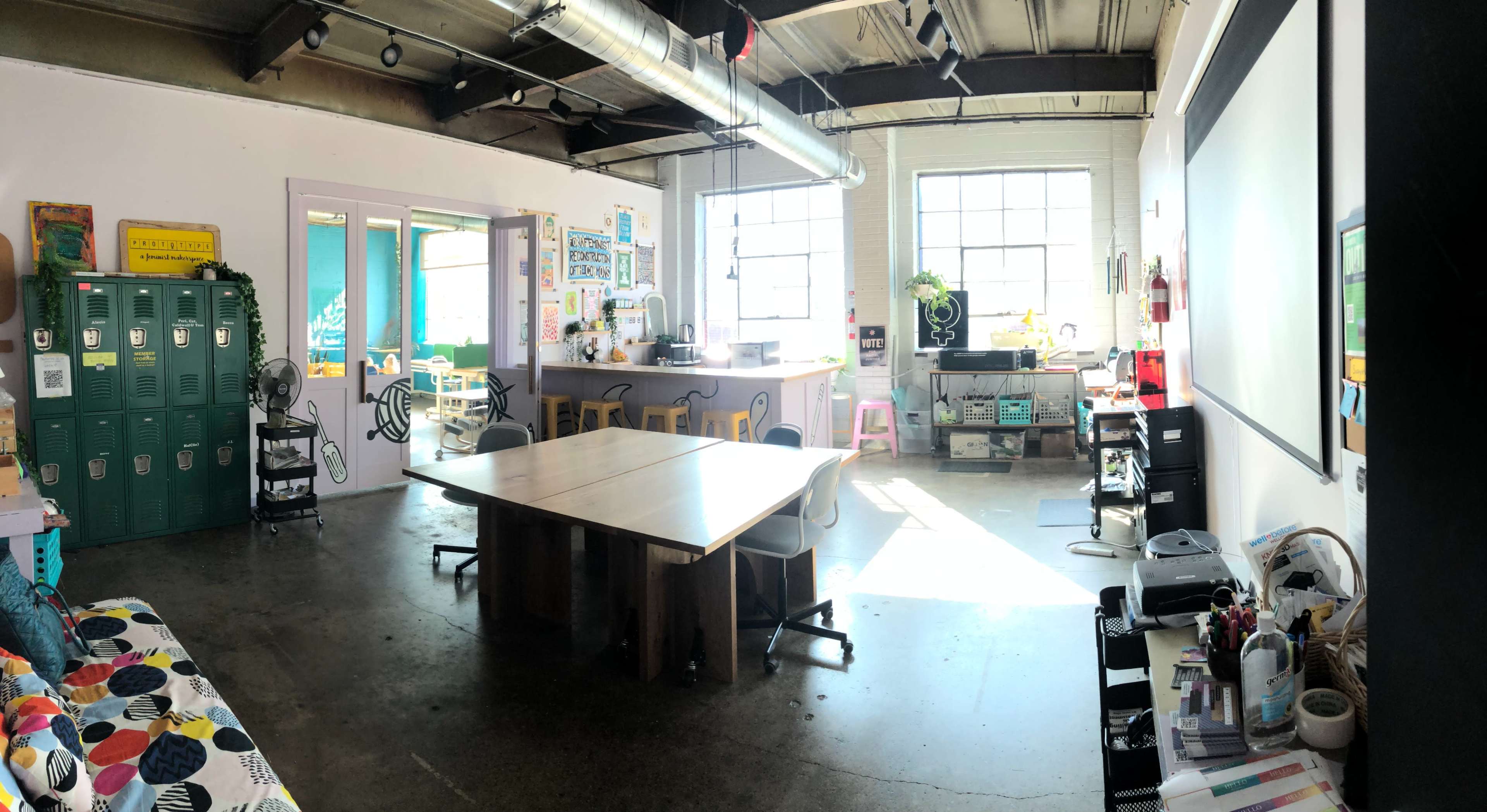 A well-lit, open workspace with a large table at the center, lockers on one side, and a kitchenette area in the background, surrounded by various creative supplies and seating.