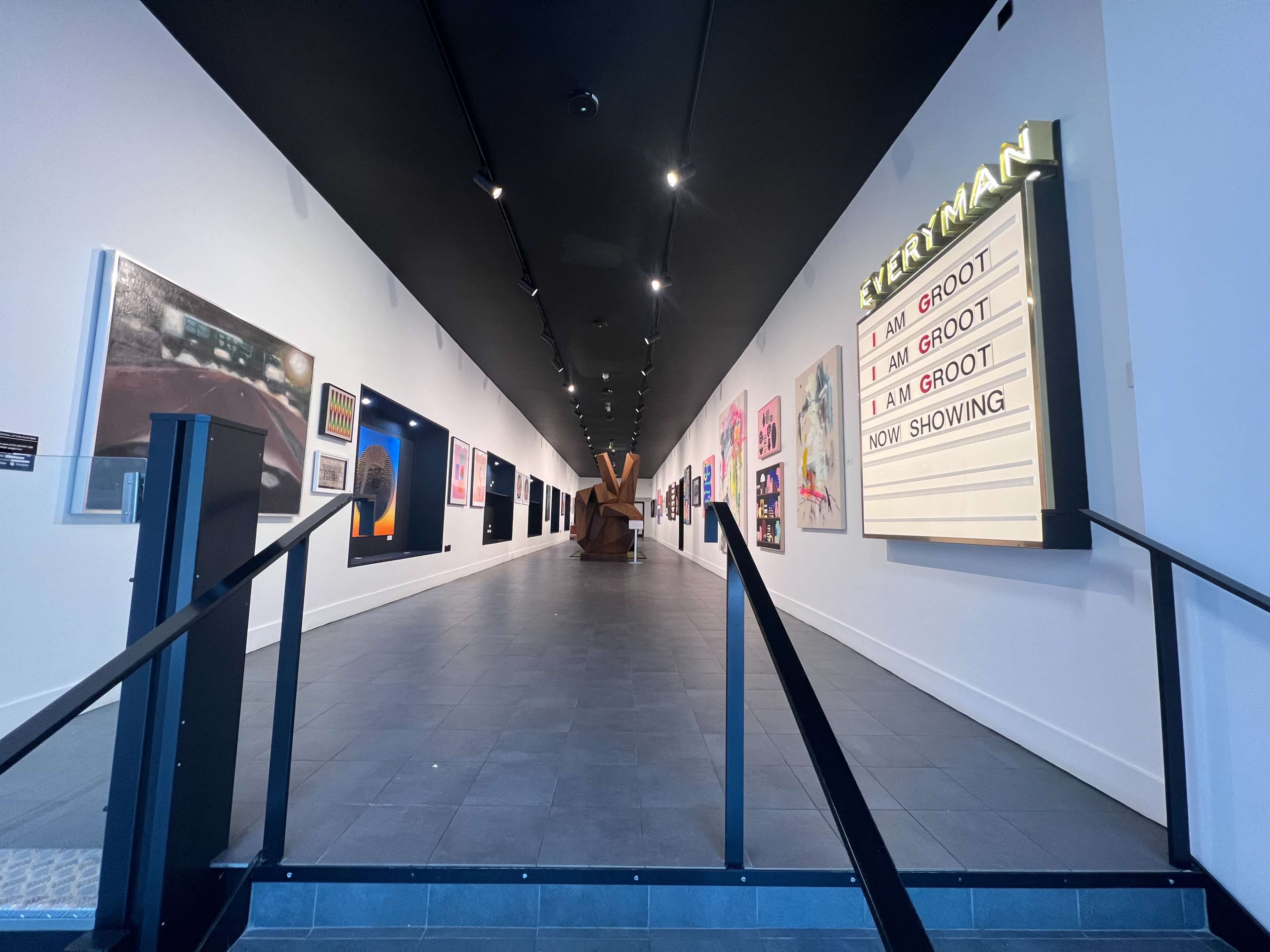 A long hallway of an art gallery featuring various artworks on display alongside a marquee sign that reads "ETERMAN" with repeated phrases "I AM GROOT."