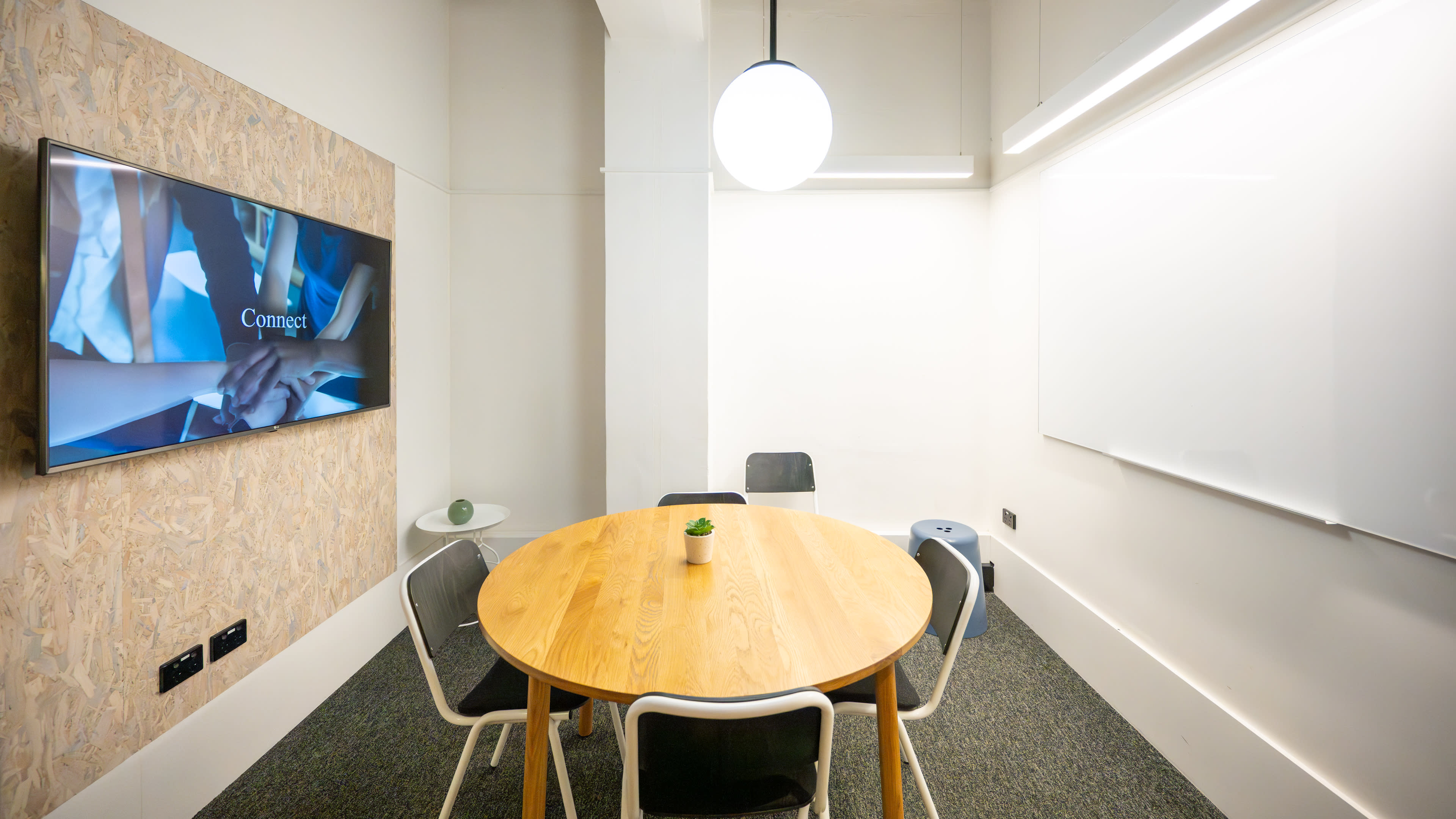 A small conference room features a round wooden table surrounded by black and white chairs, a wall-mounted screen displaying a presentation, and a whiteboard on one side.