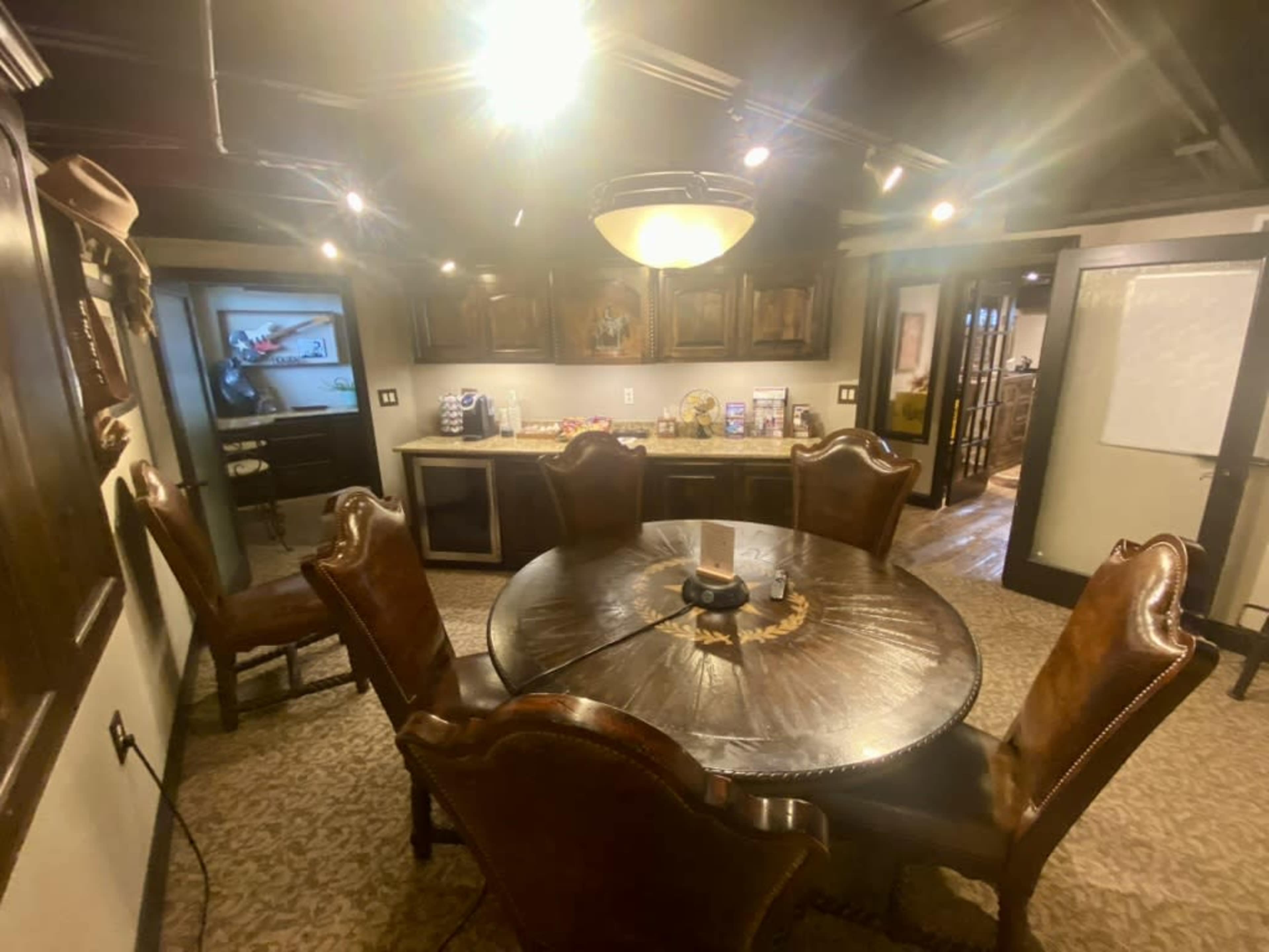 The image shows a cozy dining area with a round wooden table surrounded by leather chairs, and a kitchenette visible in the background.