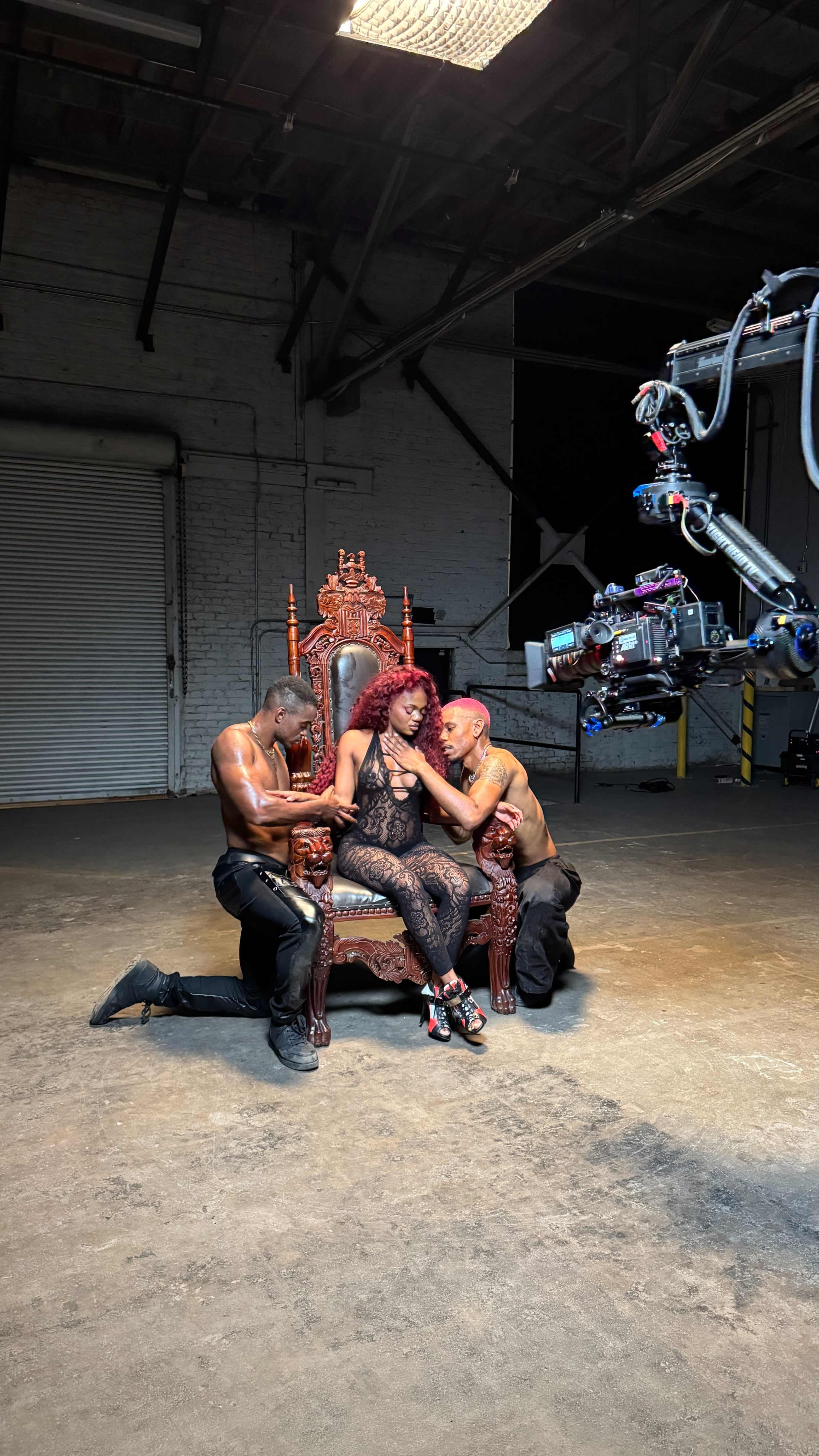 The image shows a woman seated on an ornate throne, flanked by two shirtless men kneeling beside her in an industrial space.