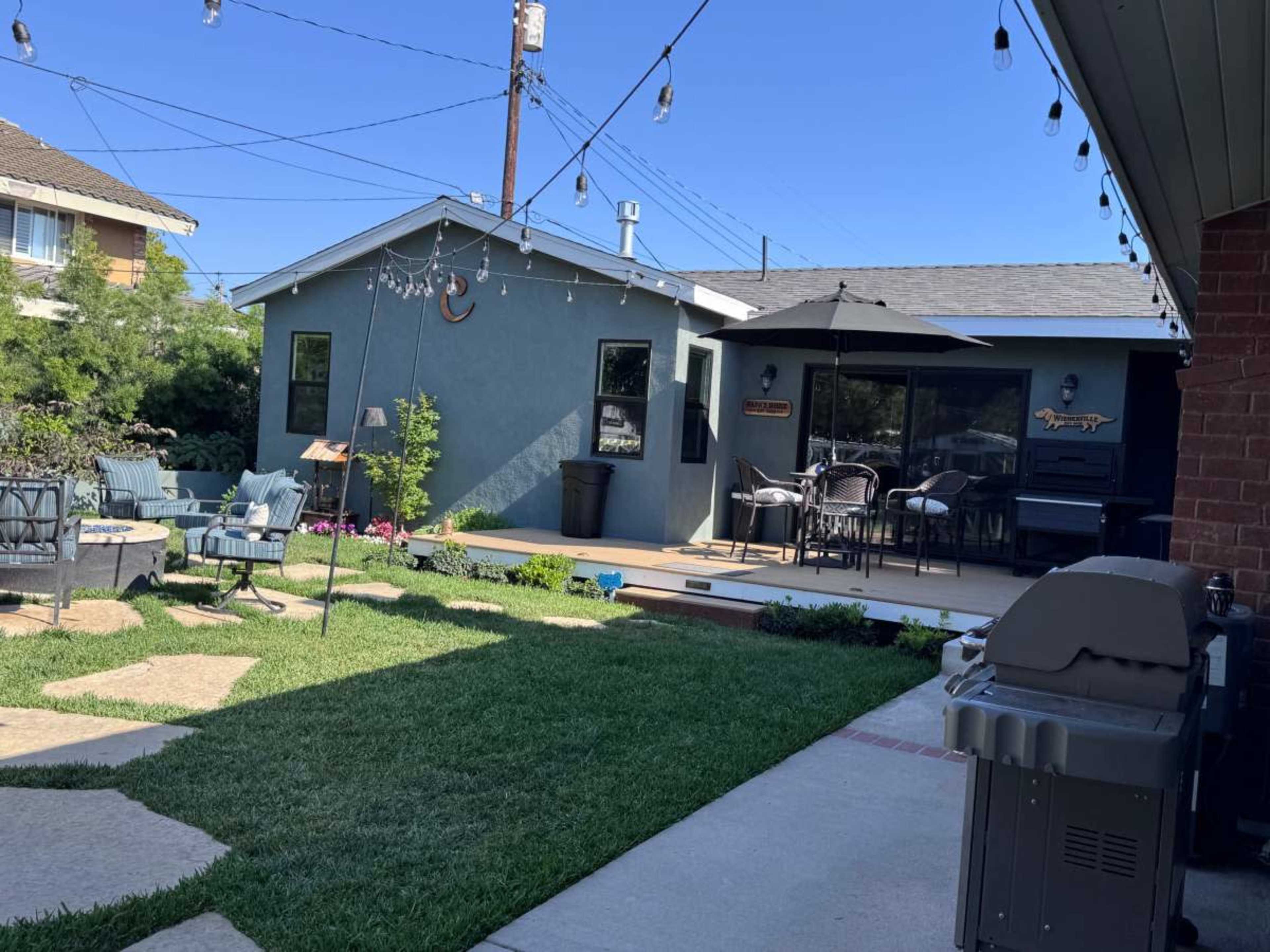 The image shows a landscaped backyard with a patio featuring outdoor furniture, a grill, and decorative string lights.