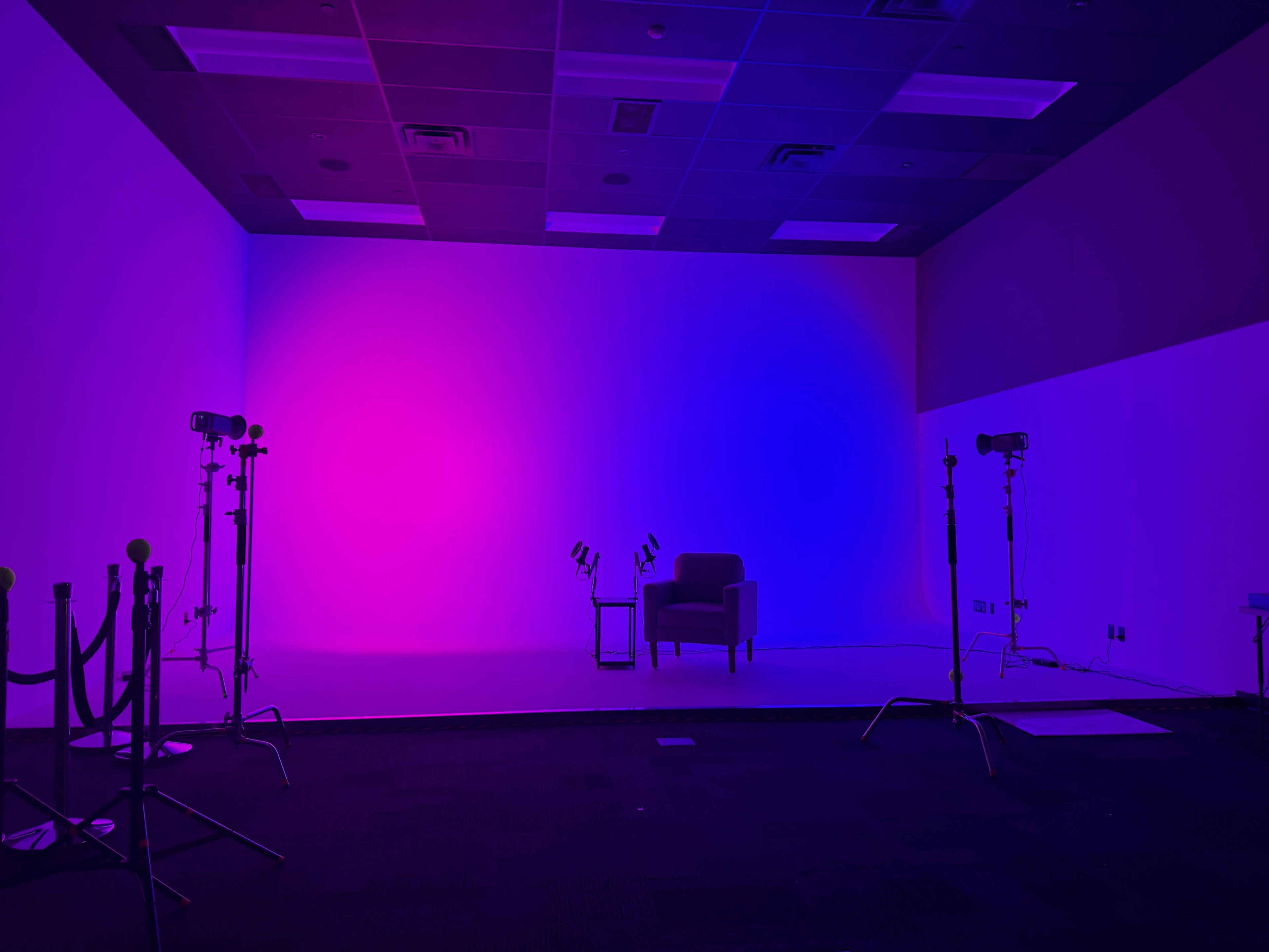 The image shows a brightly lit studio space featuring a purple and blue gradient backdrop, an empty chair, and two lighting stands on either side.